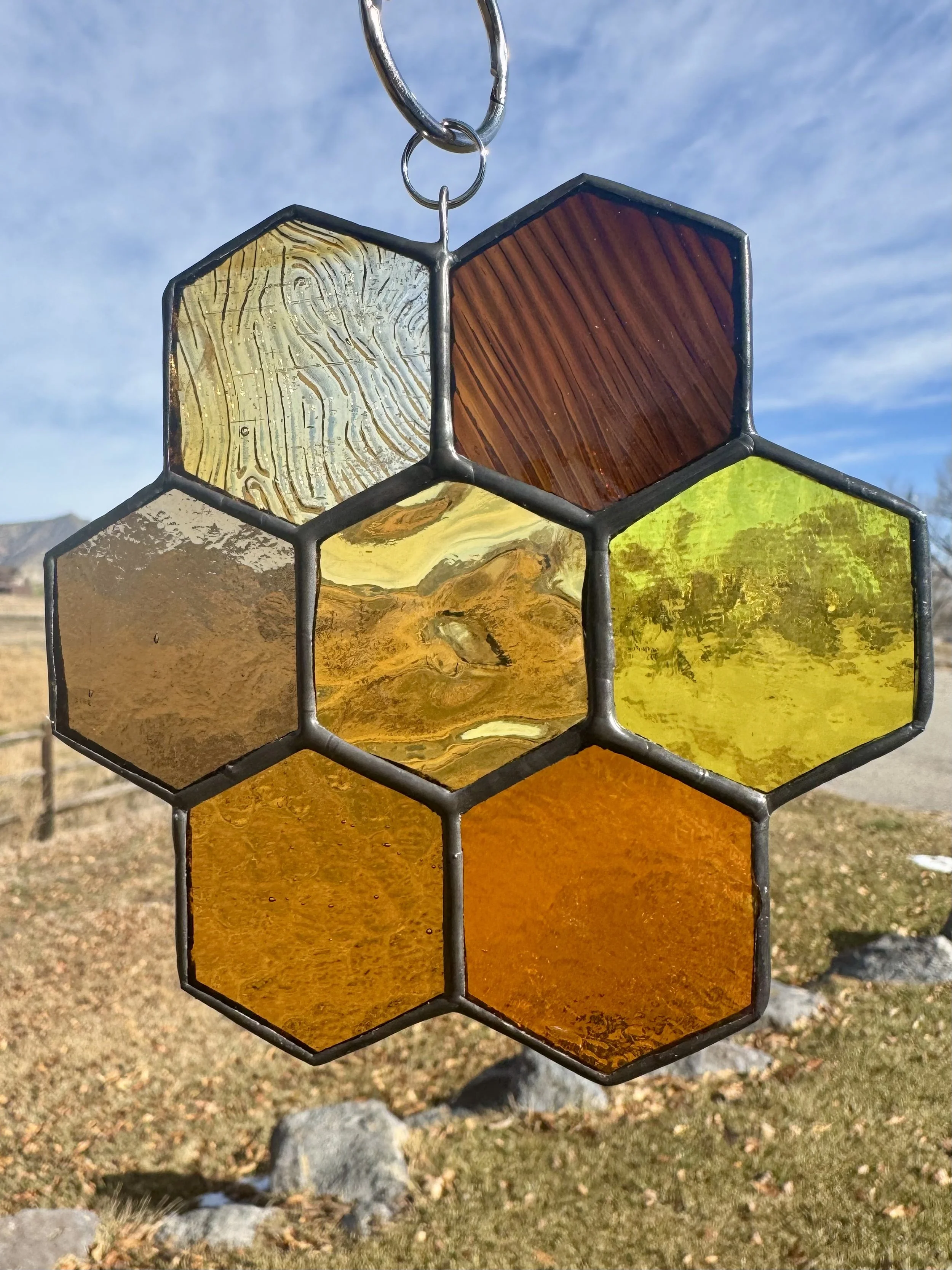 A decorative stained glass wind chime with hexagon-shaped glass pieces hanging outside, against a partly cloudy sky and landscape with rocks and grass.