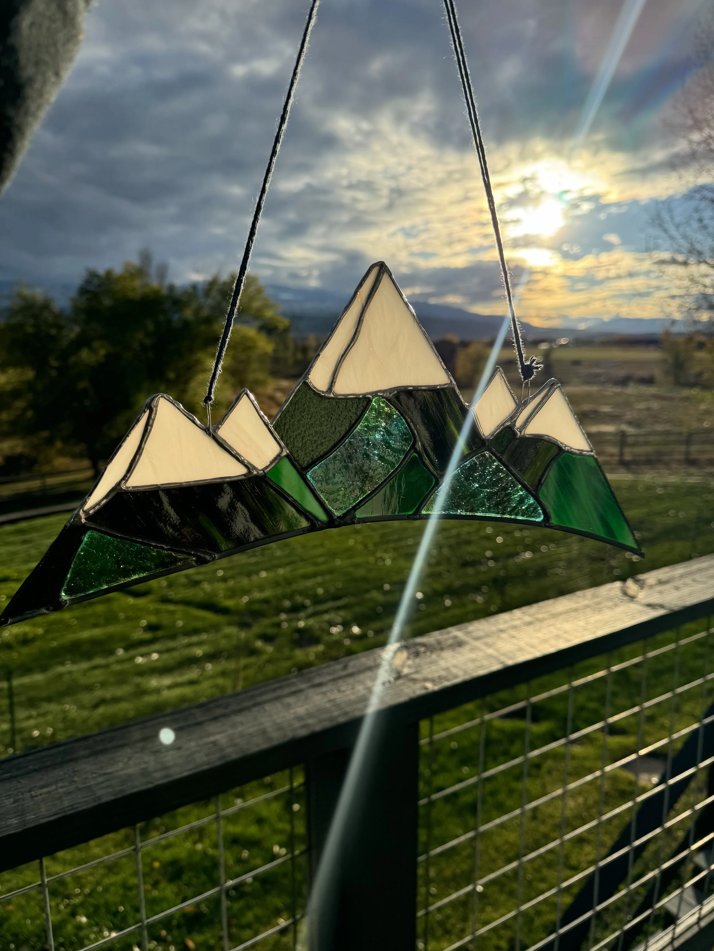 Stained glass mountain scene hanging on a wire with a cloudy sky and setting sun in the background.