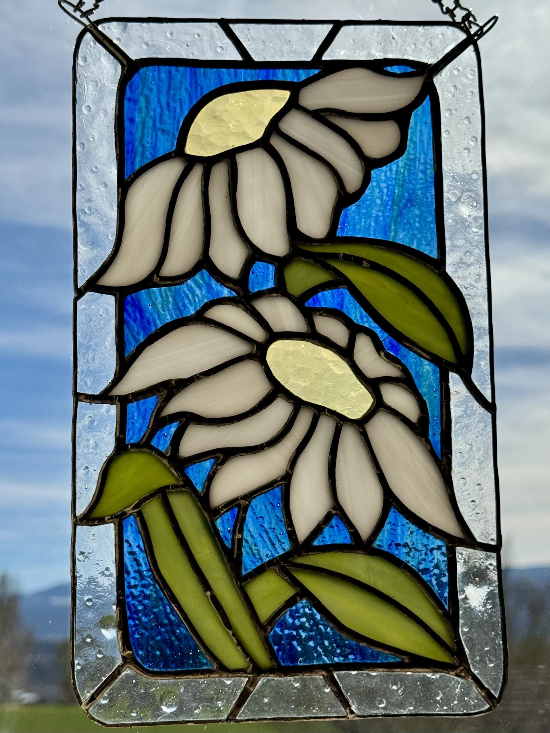 Stained glass window featuring white and yellow daffodil flowers with green stems, set against a blue background, bordered by clear textured glass.