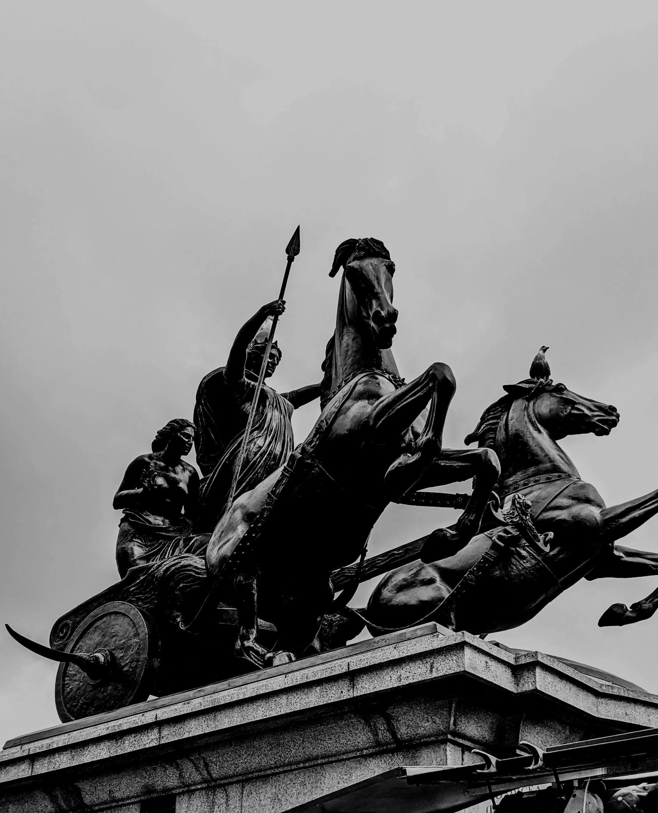 Black and white photo of a monument depicting a chariot with two horses, and two human figures, one holding a spear, on a stone pedestal.