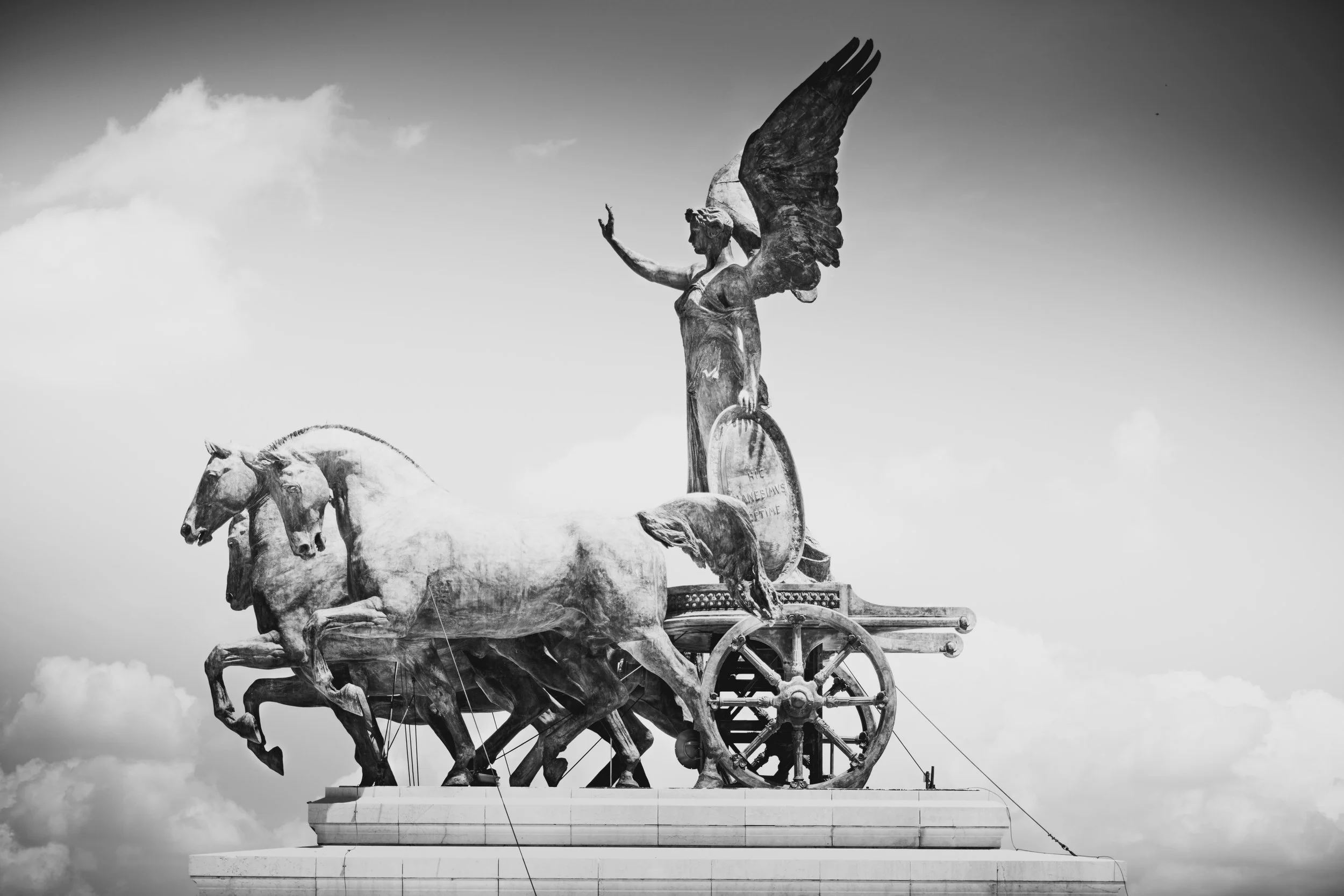 Black and white photo of a monument with three horses pulling a chariot, driven by a woman with wings, holding a circular object, all on a raised platform with a cloudy sky in the background.