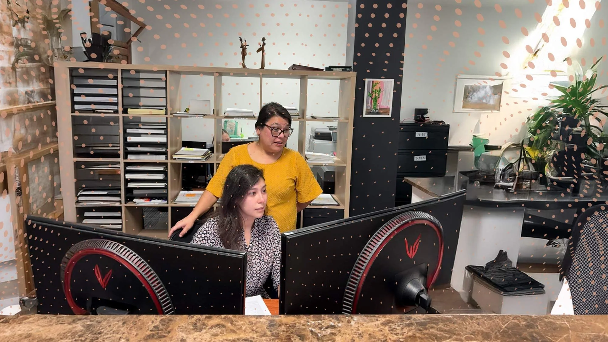 Two women working at a desk with computer monitors in an office setting, surrounded by shelves and office supplies.