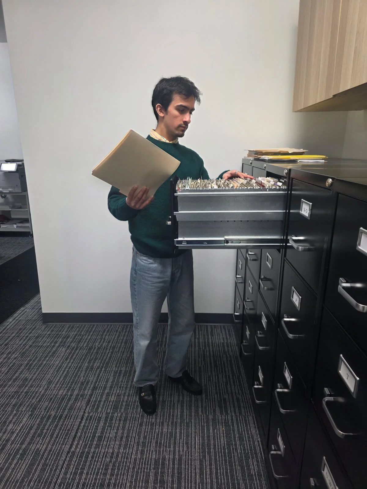 A man in business casual attire standing in an office environment, looking into a filing cabinet drawer filled with hanging file folders, holding a file folder in his left hand, and standing next to a row of filing cabinets.