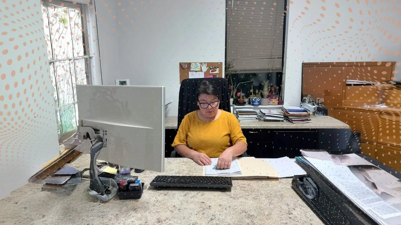 A woman working at her desk in an office, wearing a yellow shirt and glasses, surrounded by papers, a computer monitor, and various office supplies.