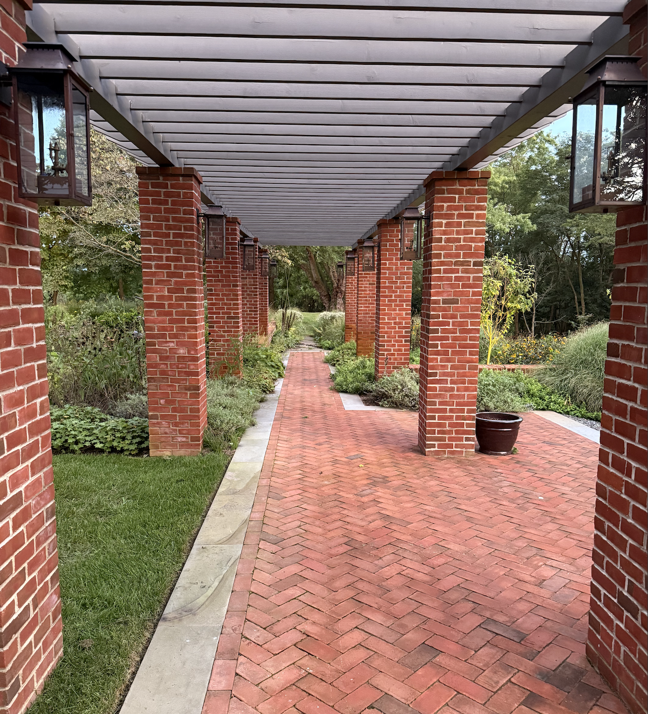 A brick walkway under a covered patio with brick columns and hanging lanterns, surrounded by lush greenery and garden plants.