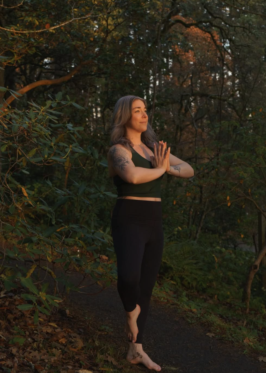 A woman practicing yoga outdoors on a trail in a wooded area during sunset, standing in a prayer position with eyes closed.
