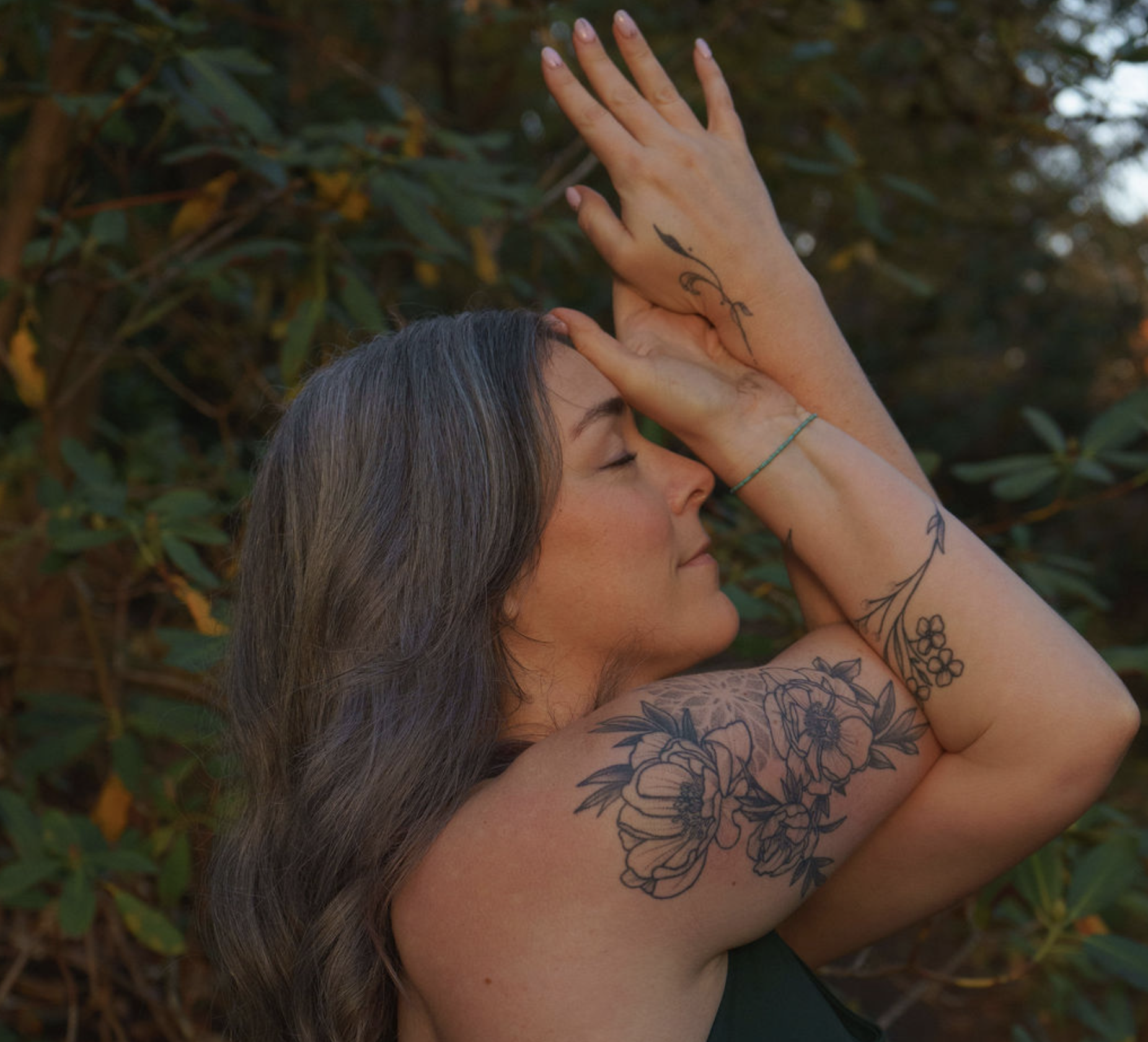 A woman with gray hair and floral tattoos on her arm and shoulder, eyes closed, smiling softly with her hands raised near her face in a peaceful outdoor setting surrounded by green foliage.