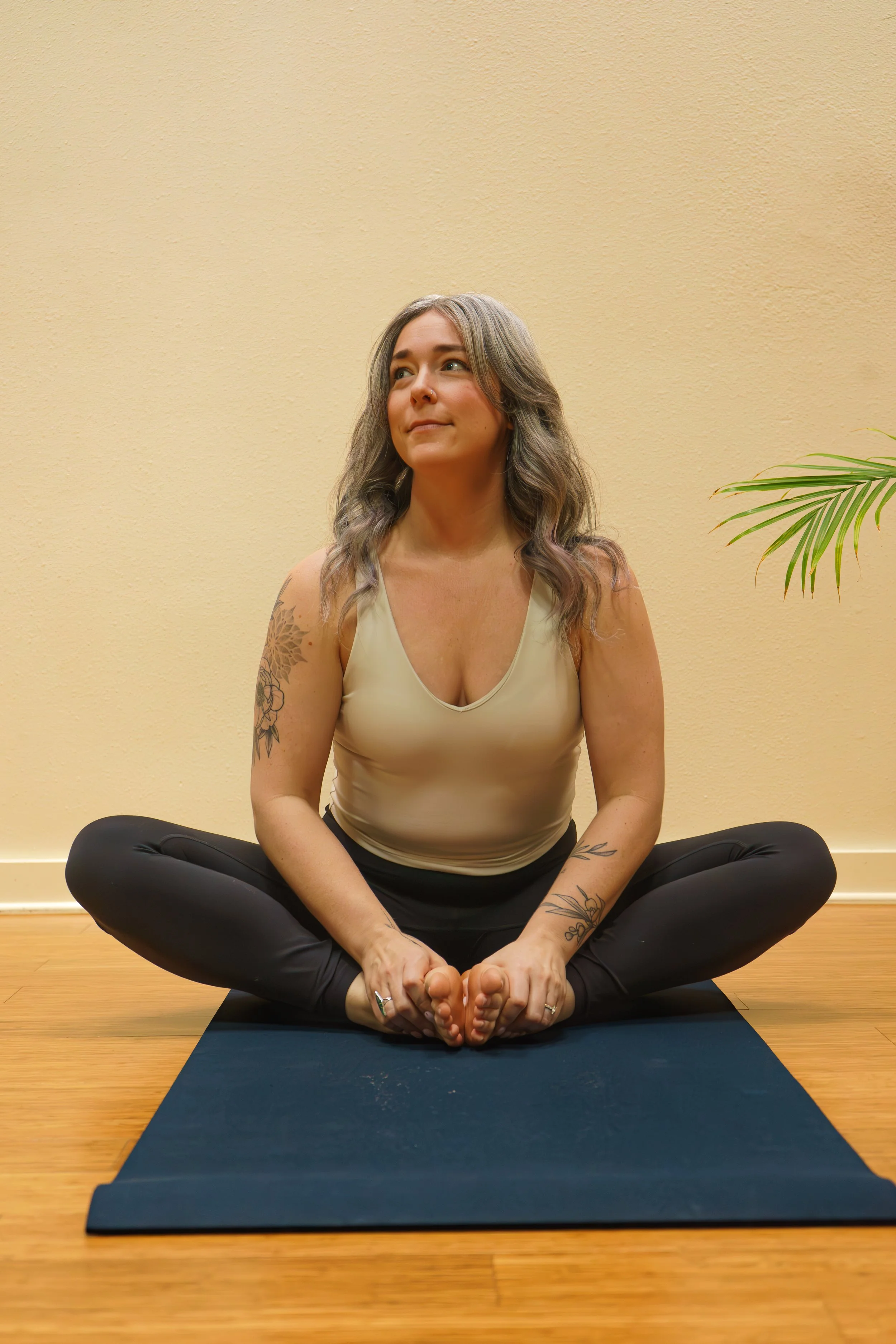 A woman with long, wavy hair sitting on a yoga mat in a cross-legged position, practicing yoga or meditation indoors with a yellow wall and a green plant in the background.