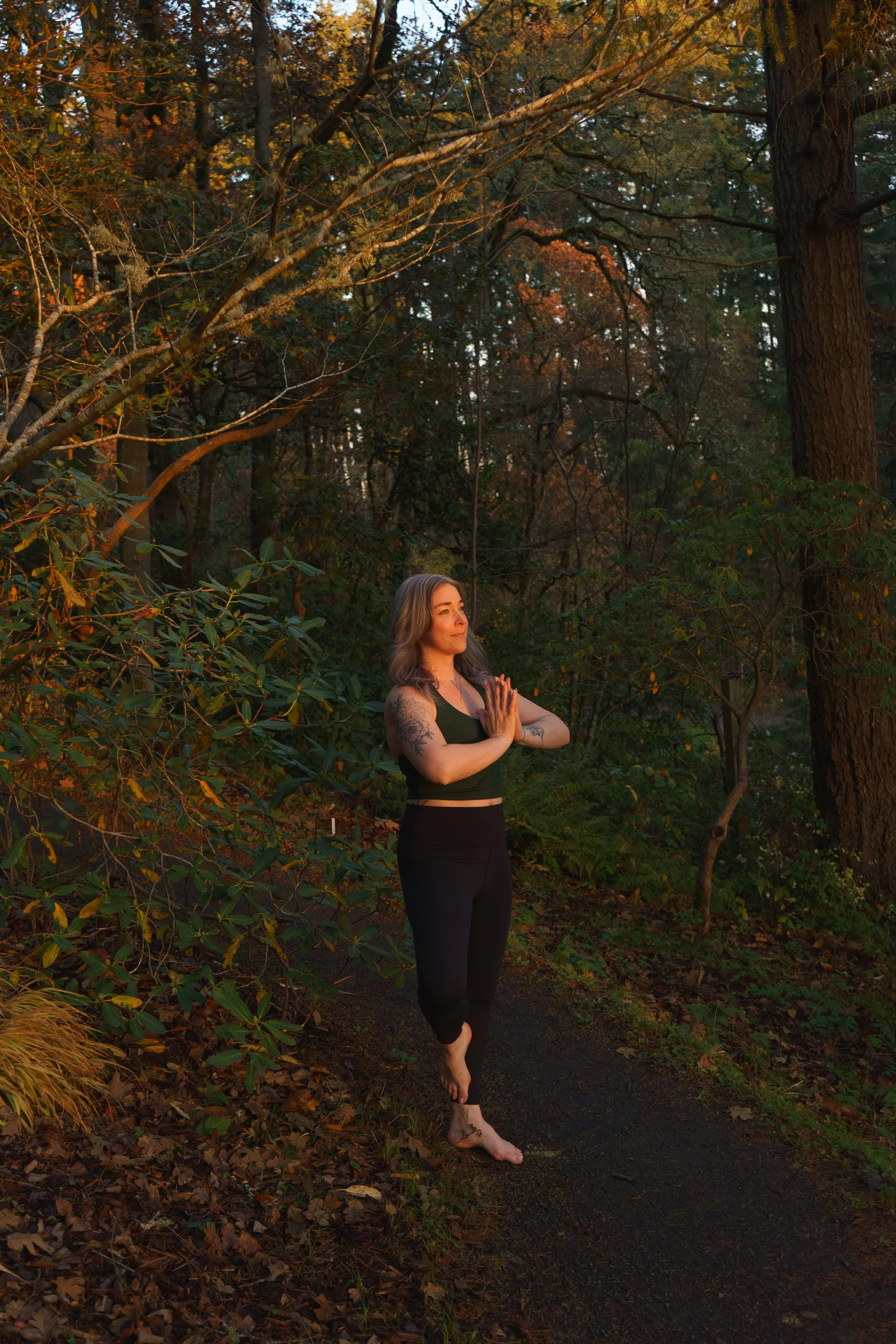 Woman practicing yoga outdoors in a forest during fall, standing on a trail with eyes closed and hands in prayer position.