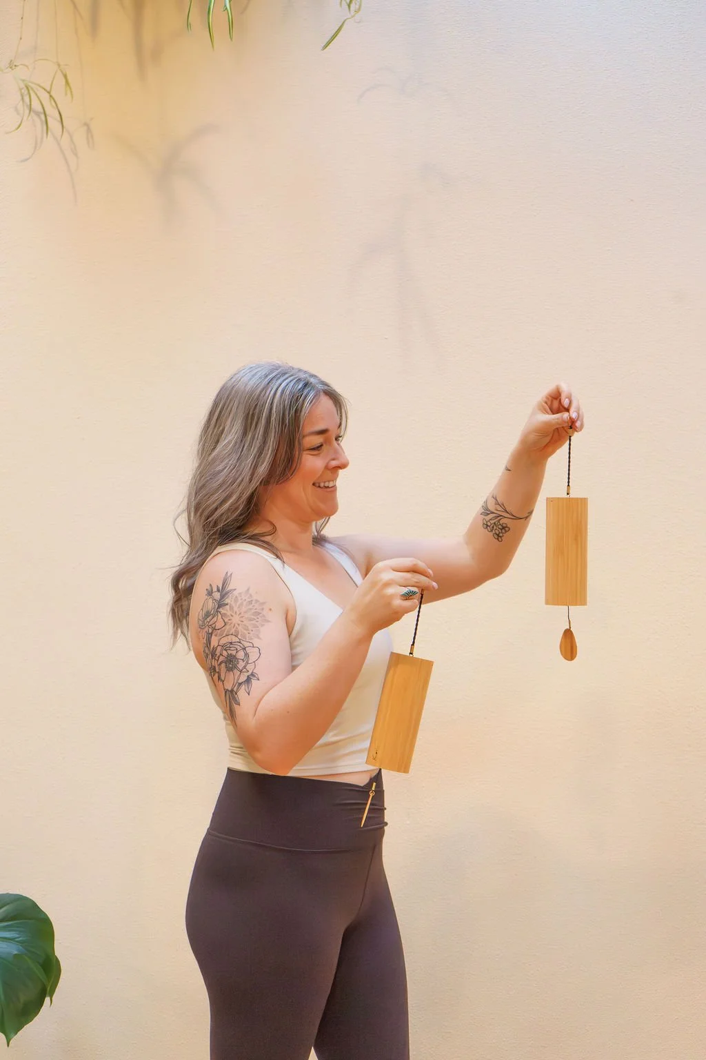 A woman with tattoos on her arm holding two wooden wind chimes with black strings, smiling, standing in front of a beige wall.
