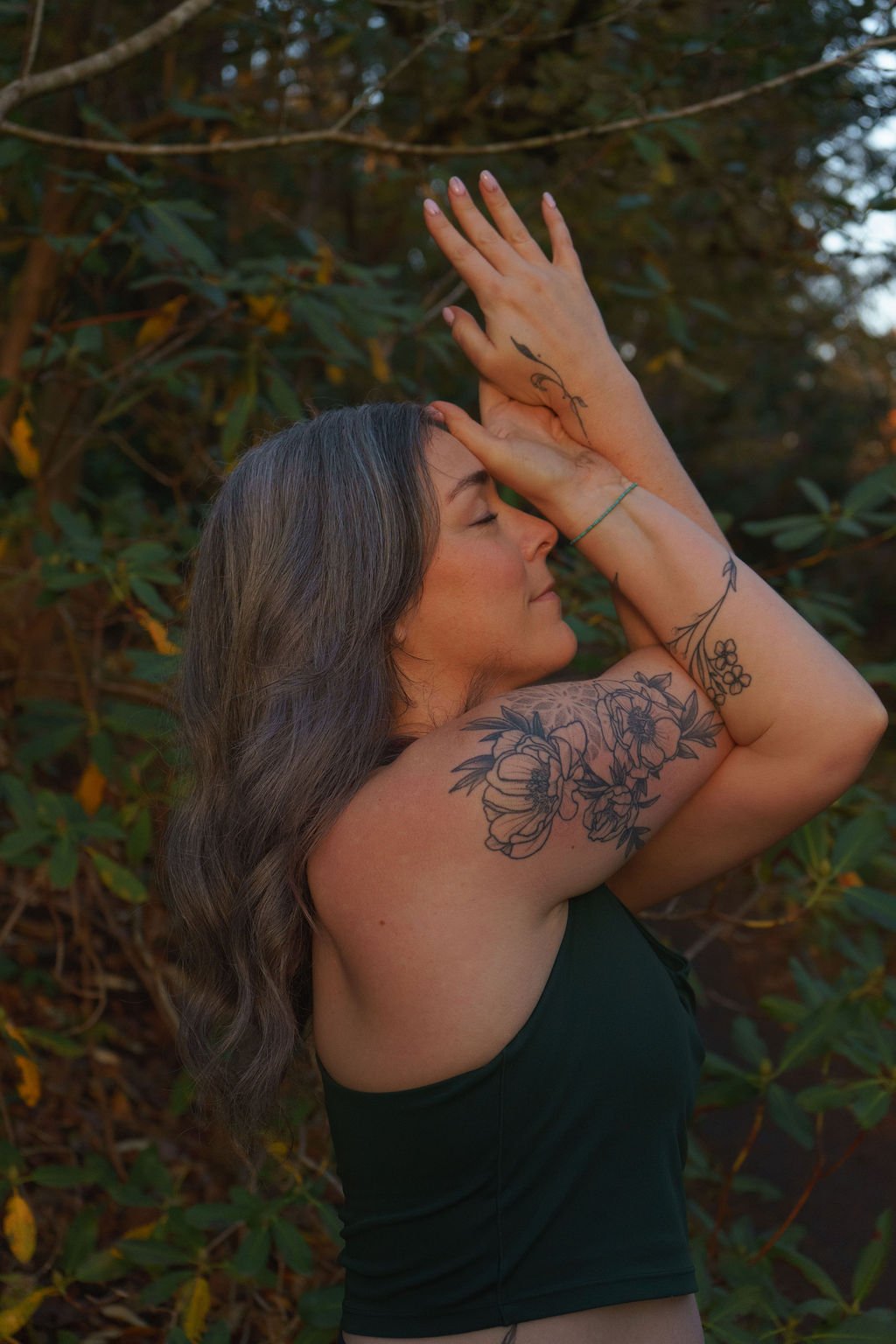 A woman with long wavy brown hair, tattoos on her arms, and wearing a black sleeveless top, posing outdoors with her eyes closed and hands raised above her head, surrounded by green foliage.