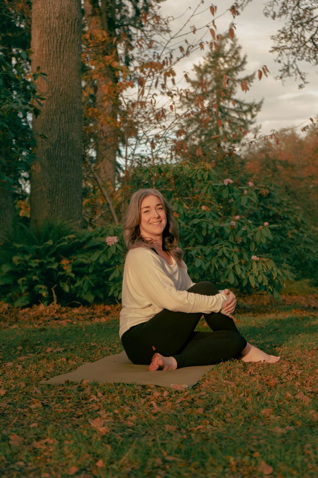 A woman with wavy hair sitting on a yoga mat outdoors in autumn, smiling, surrounded by trees and bushes with fall foliage.