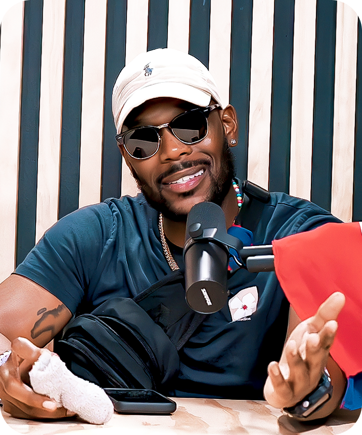 A man wearing sunglasses, a white baseball cap, and a blue shirt sits at a desk with a microphone in front of him, smiling and gesturing with his hands.