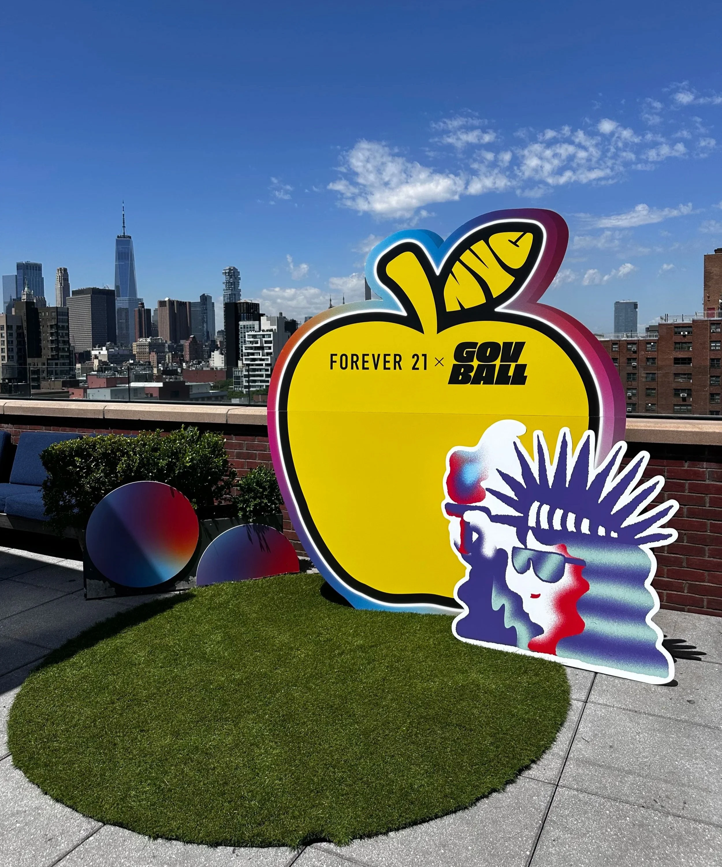 A large yellow sign in the shape of an apple with the words 'FOREVER 21 x GOW BALL' on a rooftop with a city skyline in the background. Next to the sign is a colorful cutout of a person wearing sunglasses and a headdress.
