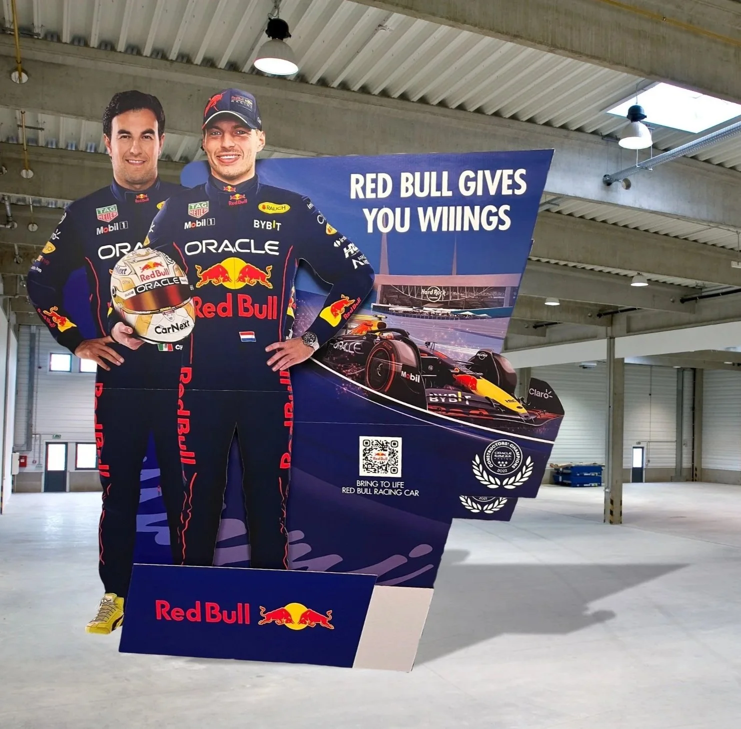 A Red Bull Racing promotional stand inside a large industrial garage, featuring two drivers in racing suits and a racing car, with the slogan "Red Bull gives you wings."