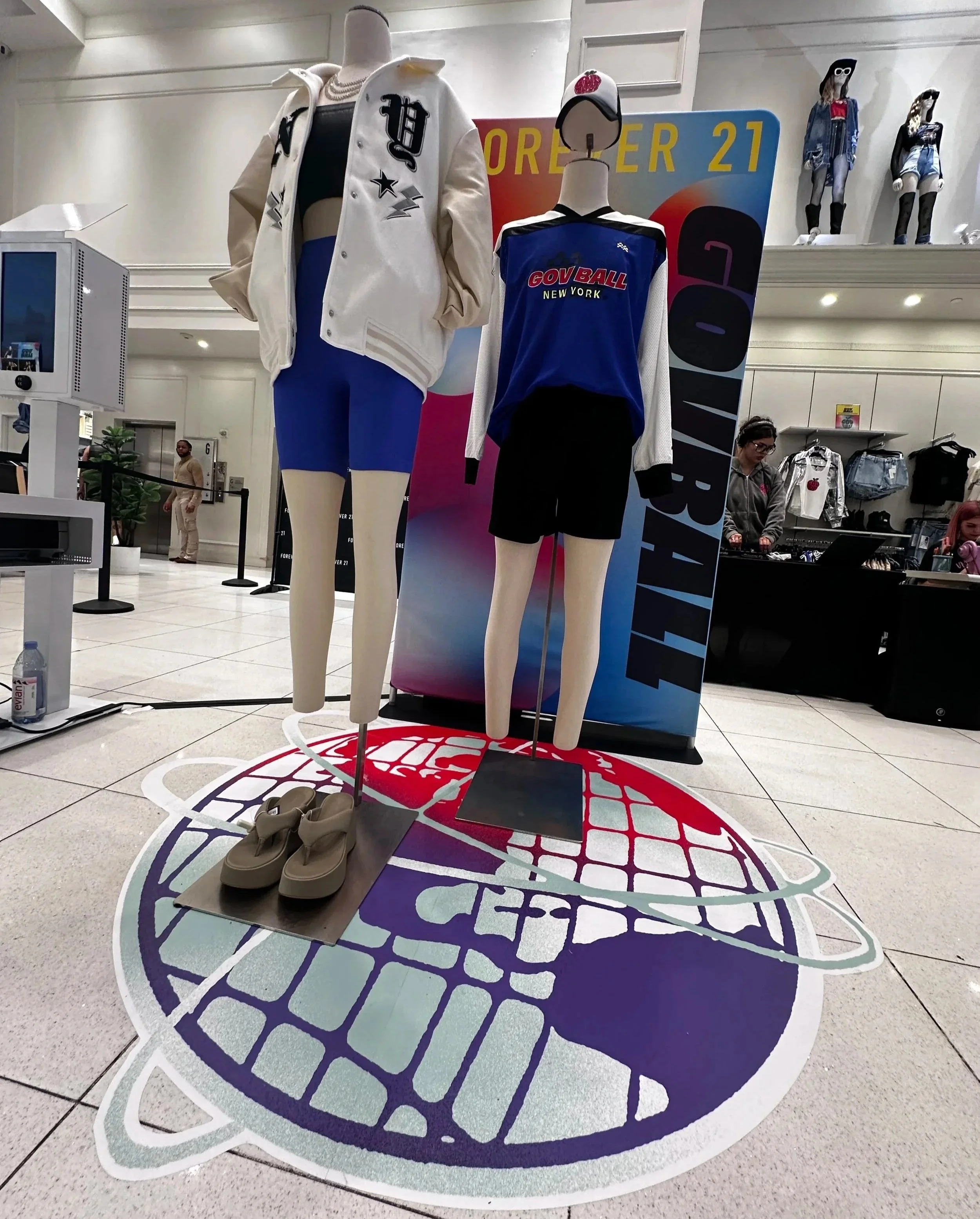 Mannequins display casual athletic outfits in a shopping mall, featuring a white jacket with black lettering and blue shorts, and a blue sports shirt with black shorts. Shoes are placed on the floor, and a promotional sign reads "ORDER 21" in the bac