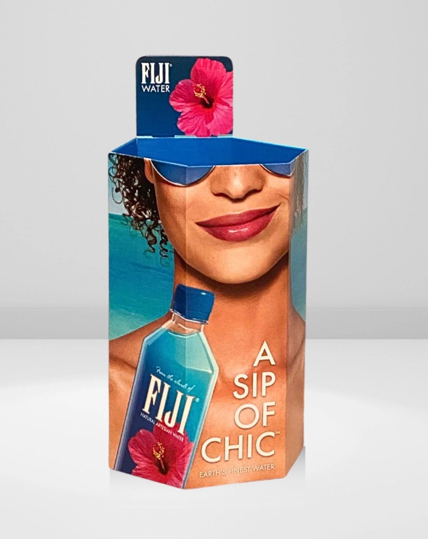 A Fiji bottled water carton featuring a woman's smiling face with sunglasses and curly hair, with text that reads 'A Sip of Chic' and a pink hibiscus flower.