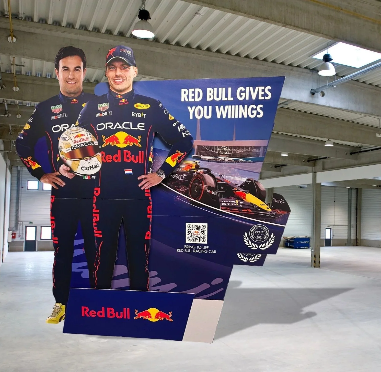 A promotional display featuring two Red Bull Racing Formula 1 drivers in racing suits, one holding a helmet, with a backdrop of a race car and the slogan "Red Bull gives you wiings." The display includes a QR code and Red Bull branding.