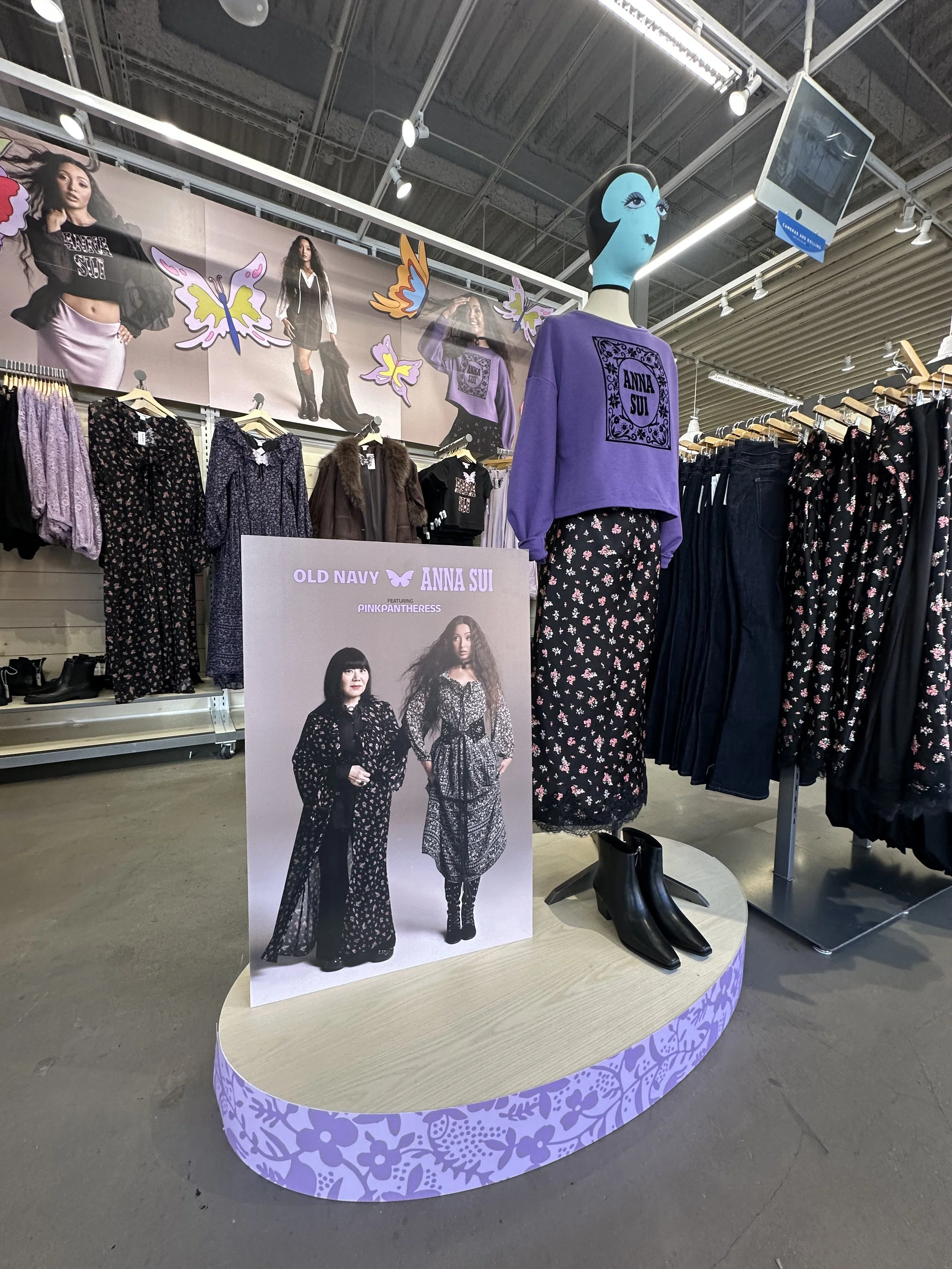 Display of women's clothing featuring Anna Sui collection, including a purple sweatshirt with black logo, floral patterned long skirt, and black ankle boots, with promotional poster and butterfly decorations.