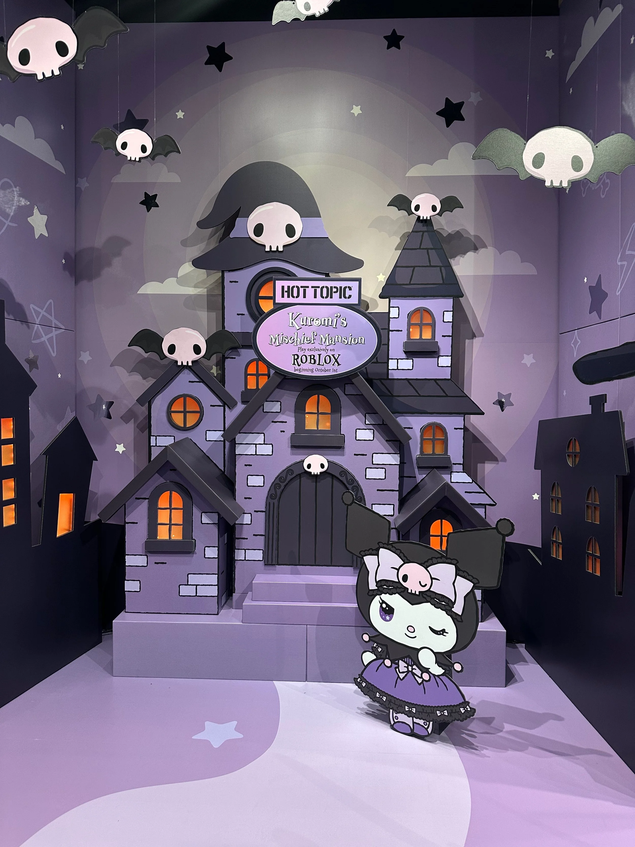 The image shows a Halloween-themed display resembling a spooky mansion in purple and black, decorated with skull and bat cutouts hanging from the ceiling and walls with glowing orange windows. In front, there is a cartoon character figure with a larg
