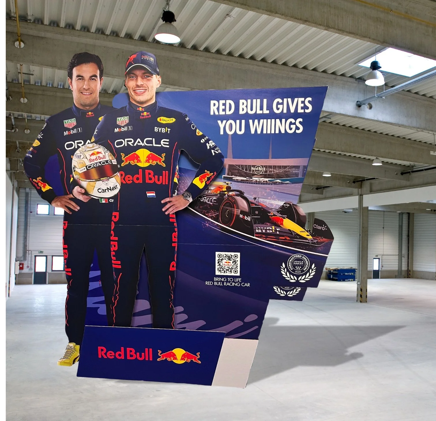 A promotional display featuring two Red Bull Racing Formula 1 drivers in racing suits, one holding a helmet, with a backdrop of a race car and the slogan "Red Bull gives you wiings." The display includes a QR code and Red Bull branding.