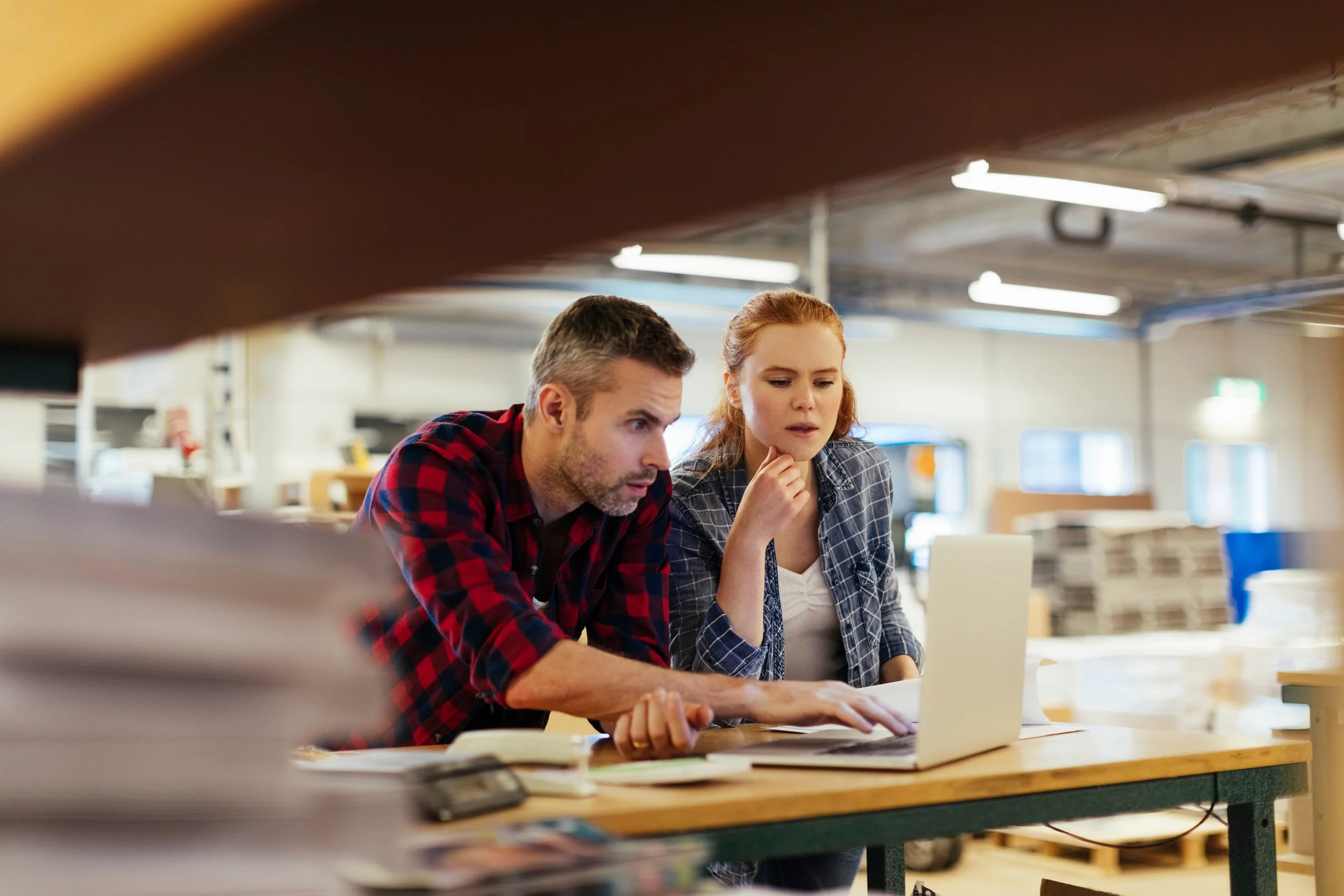 A man and woman working together on a laptop in a warehouse or industrial space, surrounded by stacks of paper and boxes.