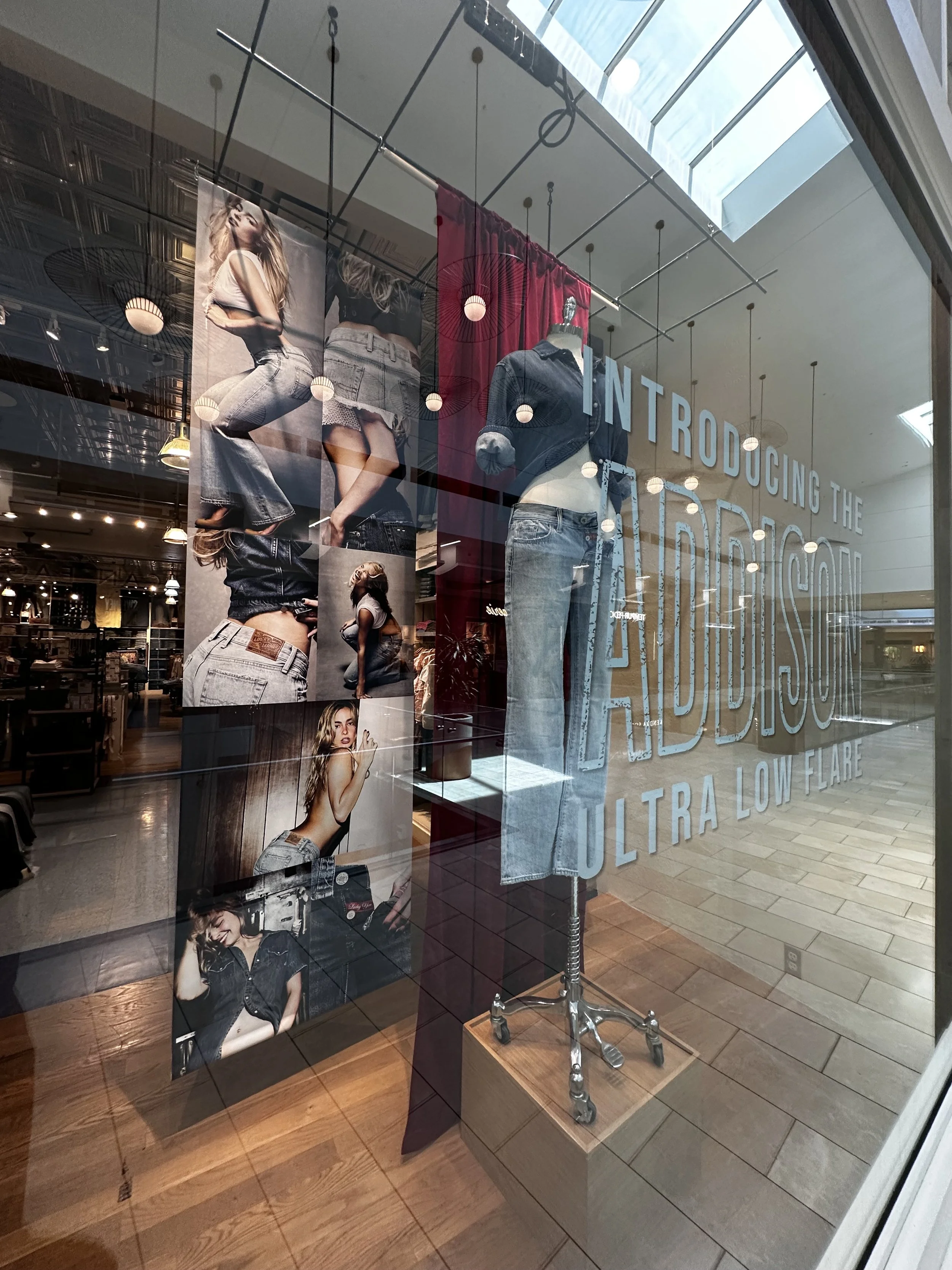 Store display window with a mannequin wearing denim jeans and a denim jacket, surrounded by fashion photos of women in denim clothing, and the text 'Introducing the Aldrich Co. Ultra Low Flare' on the glass.