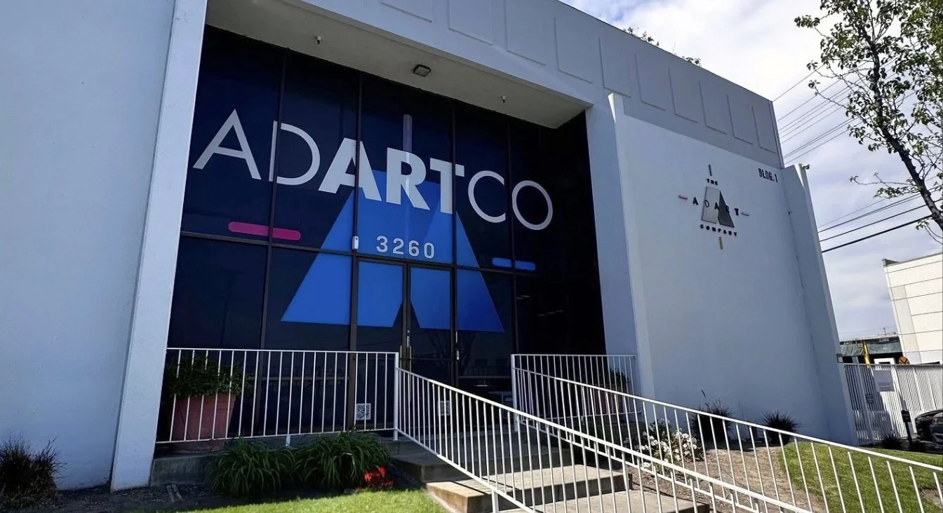 Exterior of a modern building with large glass windows displaying 'ADARTCO' and the address '3260'; white walls, a ramp with white railings, and small landscaped garden in front.