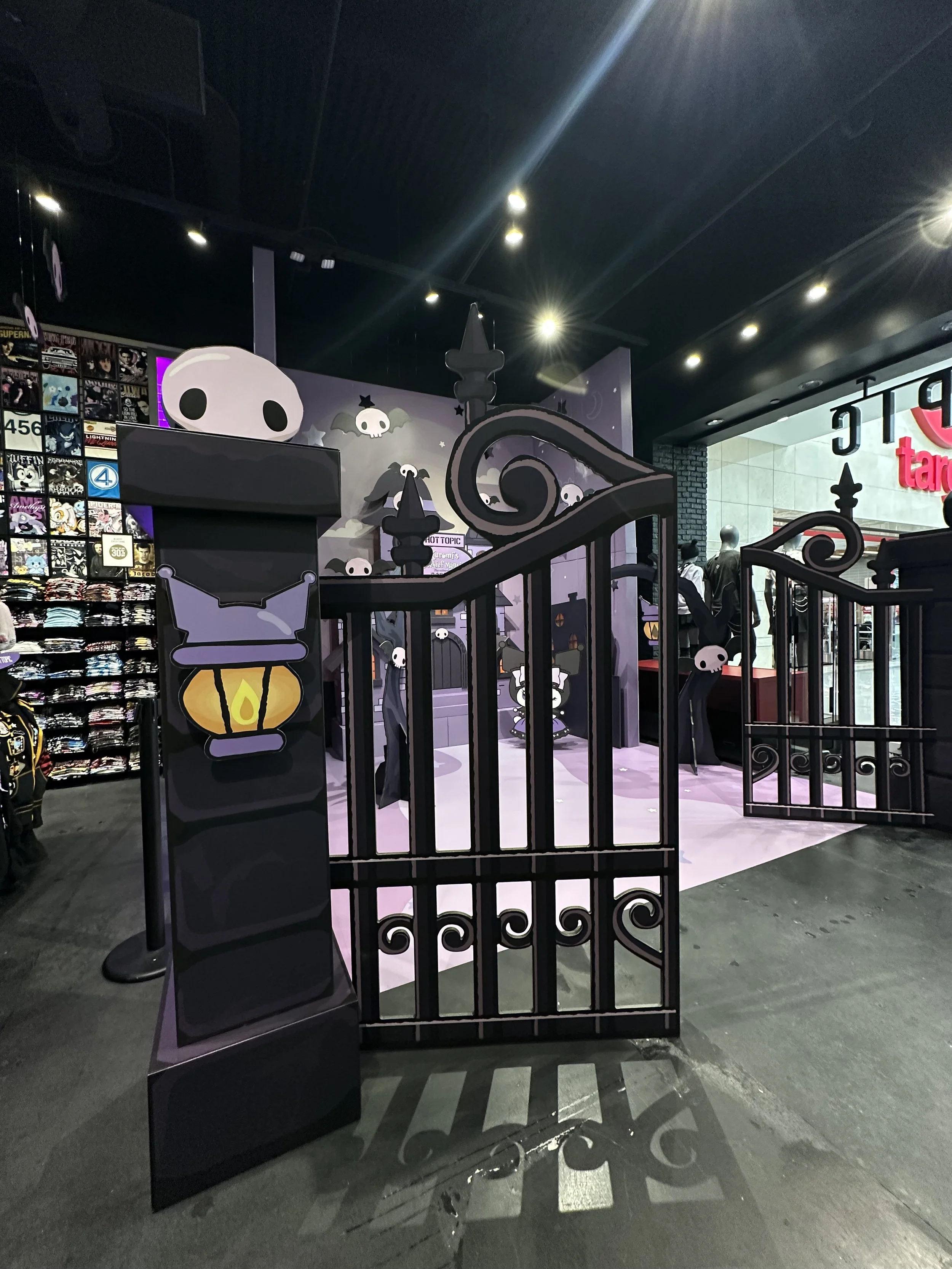 Halloween-themed display with black wrought iron fence, cartoon skeletons, and purple wall with skull illustrations inside a shopping store.