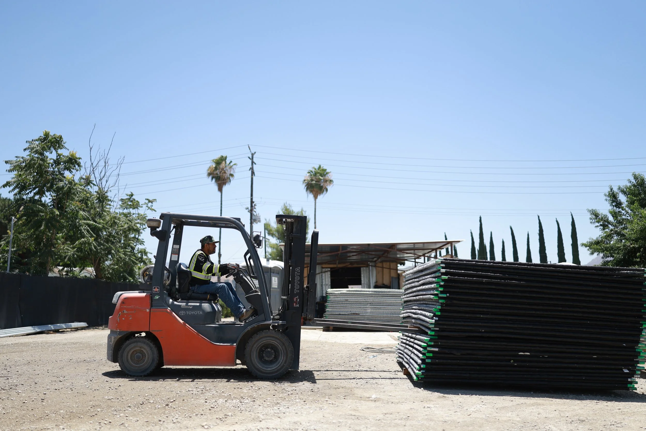 A construction worker driving a forklift at a construction site, moving black and gray pipes or materials, with trees and utility poles in the background under a clear blue sky.