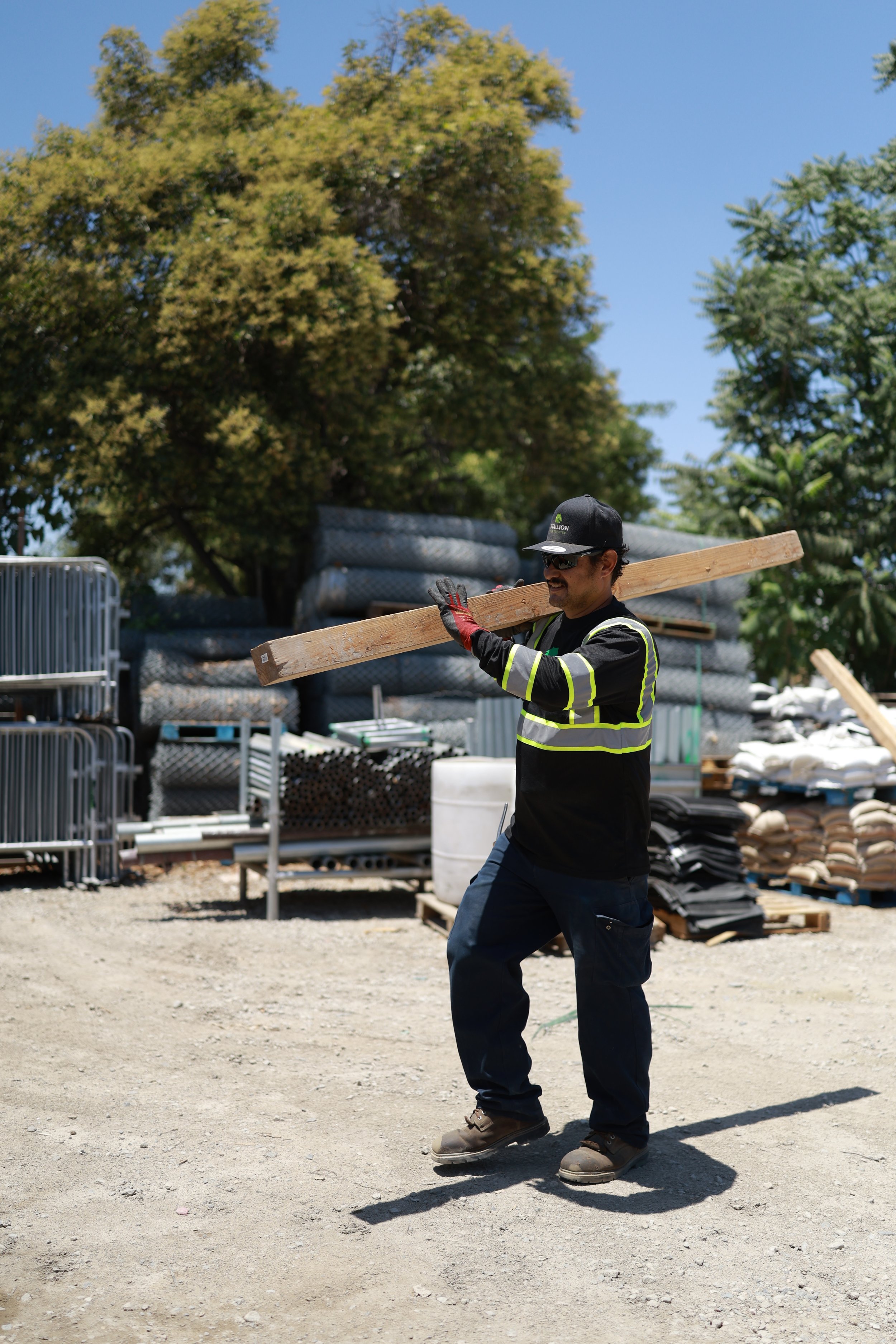 A construction worker wearing a black uniform with reflective stripes, gloves, sunglasses, and a cap, carrying a wooden plank on his shoulder at an outdoor construction site with trees and construction materials in the background.