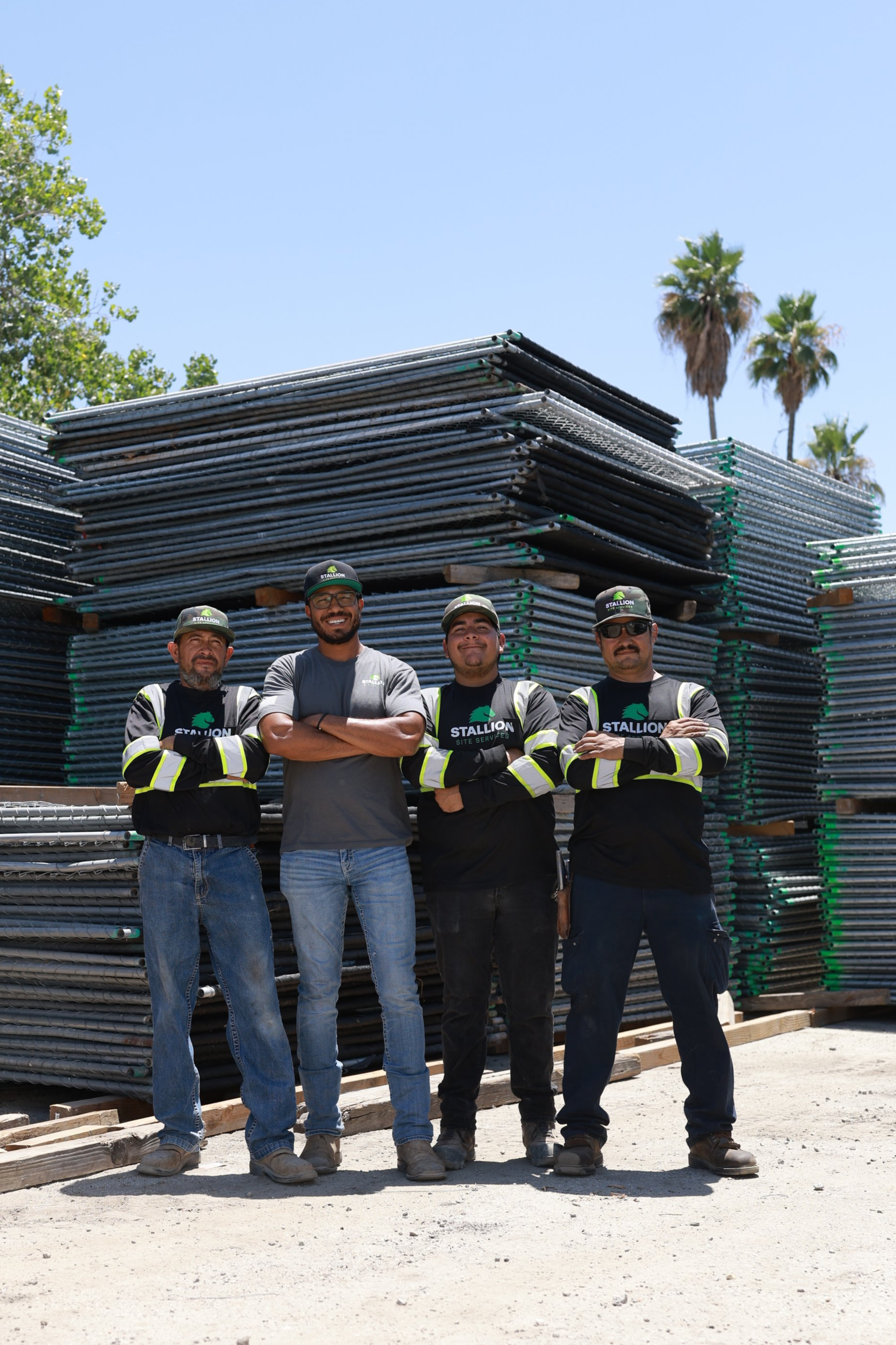 Four construction workers standing with arms crossed in front of stacked metal scaffolding on a construction site, wearing helmets and uniforms with 'STALLION' logo.
