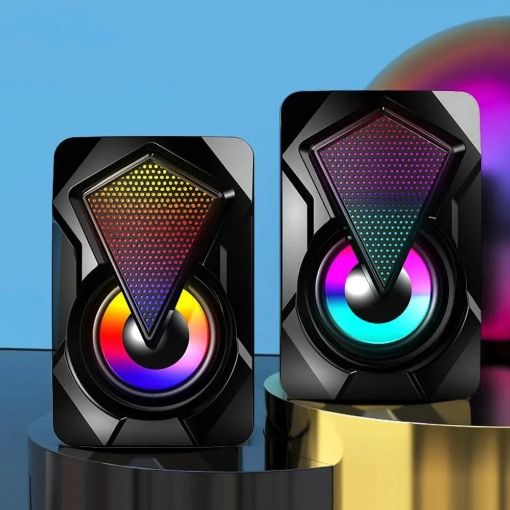 Two black computer speakers with colorful, rainbow LED lights, placed on black and gold cylindrical platforms against a gradient blue and purple background.