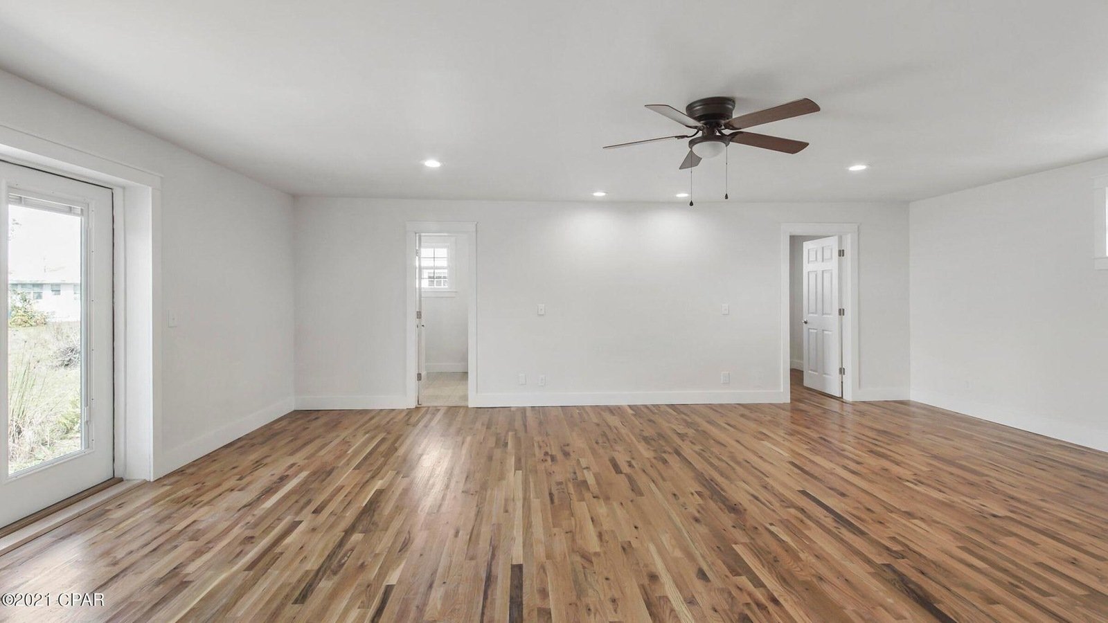 Empty living room with hardwood floors, white walls, a ceiling fan, and multiple windows, with two doors leading to other rooms.