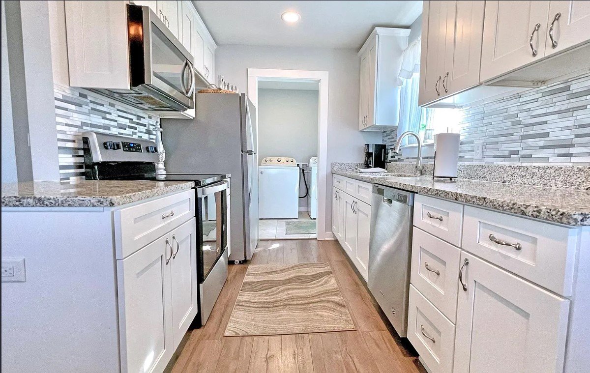 Well-lit kitchen with white cabinets, granite countertops, stainless steel appliances, and a mosaic tile backsplash. A rug is on the wood floor, with a laundry room visible through an open doorway at the end.