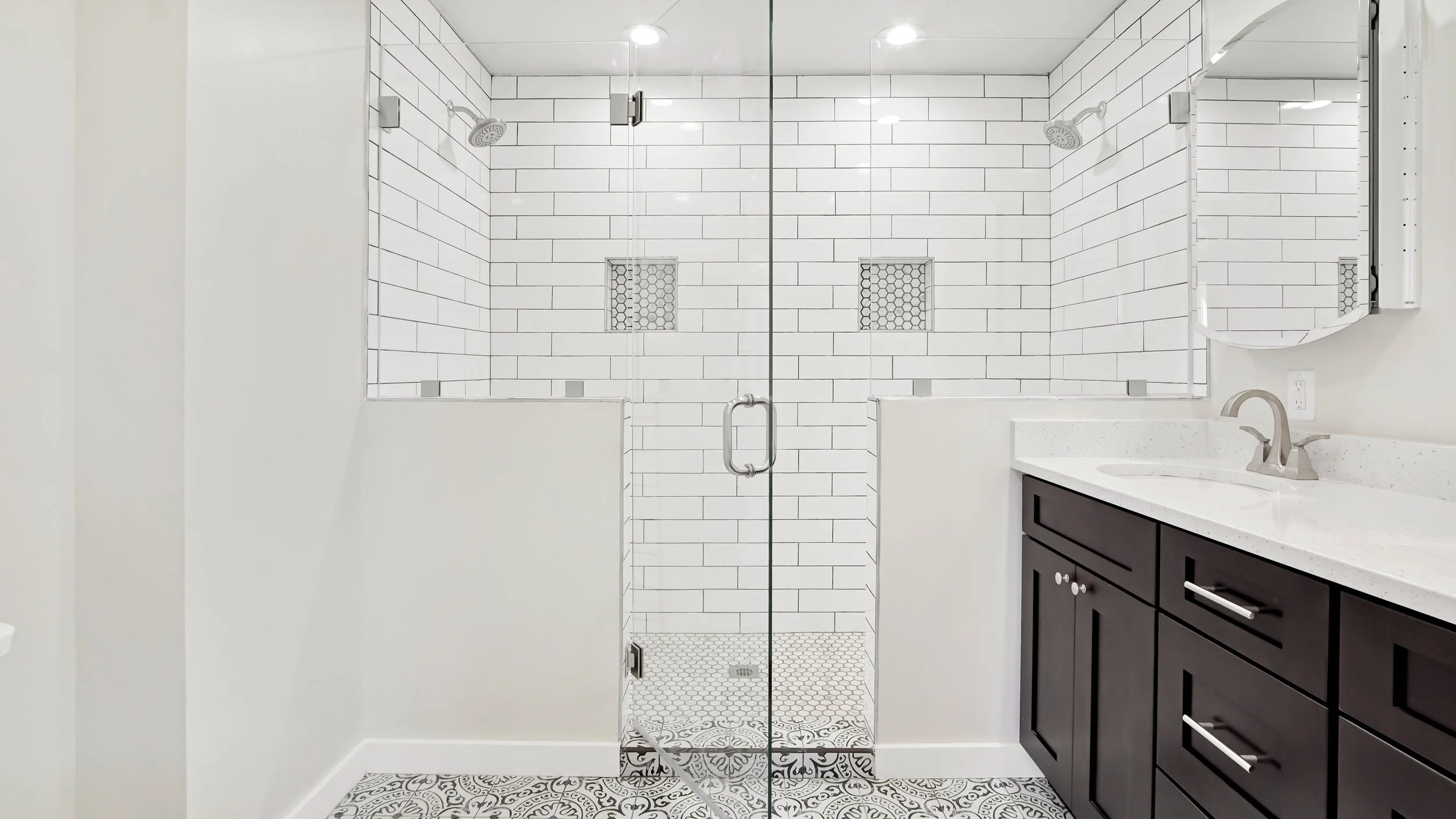 Modern bathroom with a glass shower enclosure, white subway tile walls, decorative floor tiles, and a dark wood vanity with a light countertop and a round mirror.