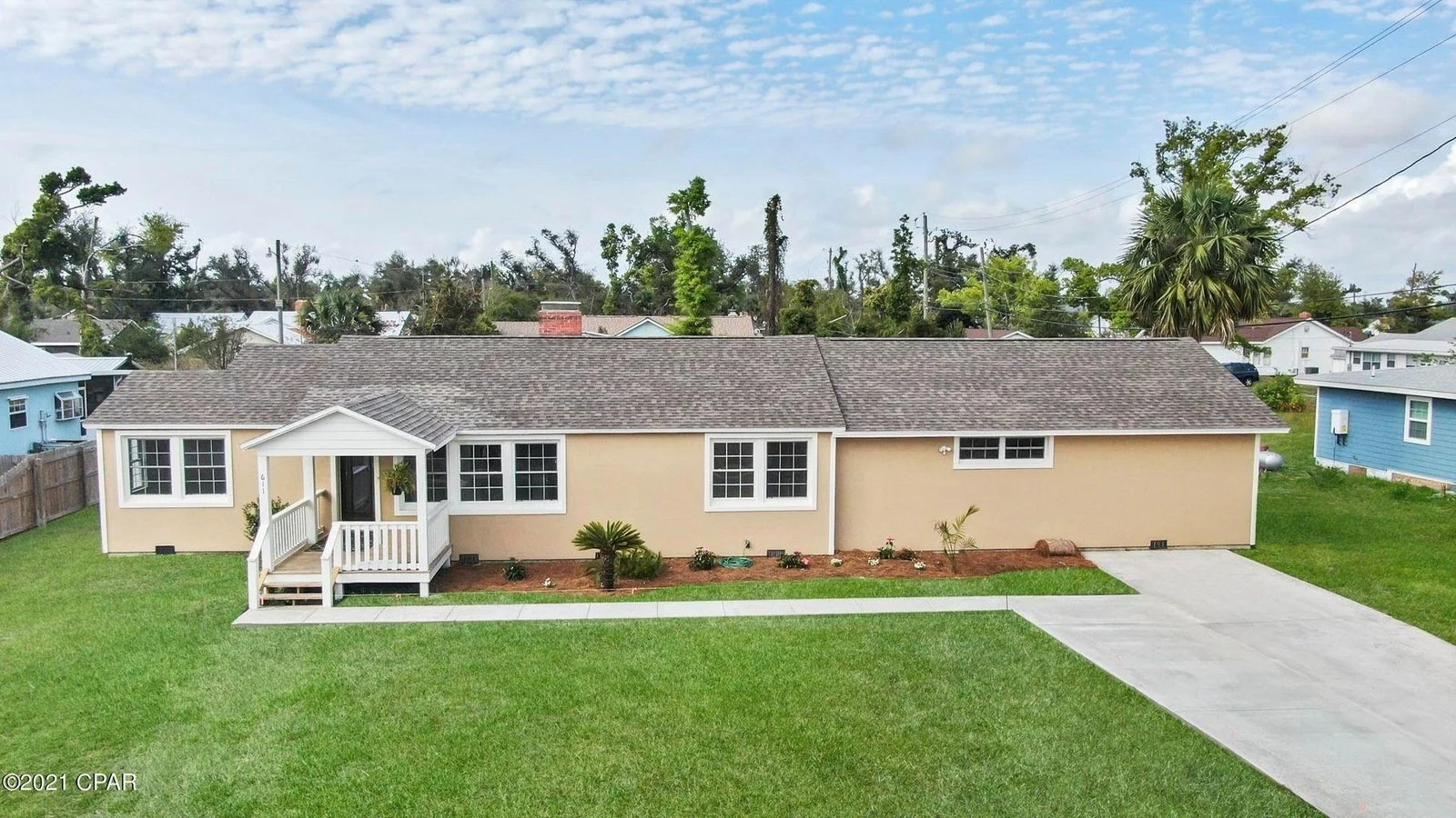 A single-story house with a beige exterior, white trim, and a small front porch, situated in a grassy yard with a concrete driveway on the right side.