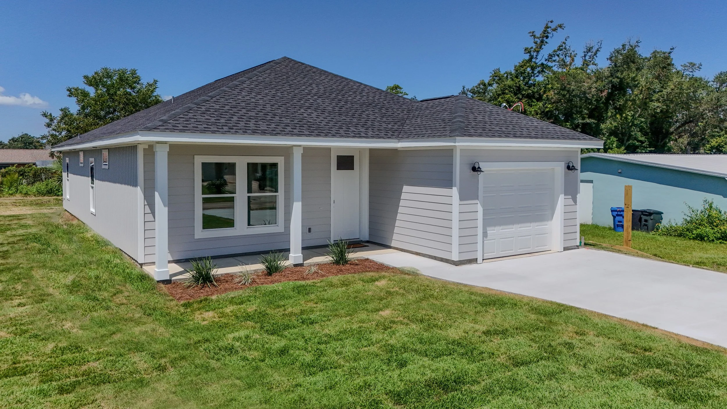 Front view of a white house with a gray shingled roof, front porch, and attached garage, surrounded by a green lawn and trees.