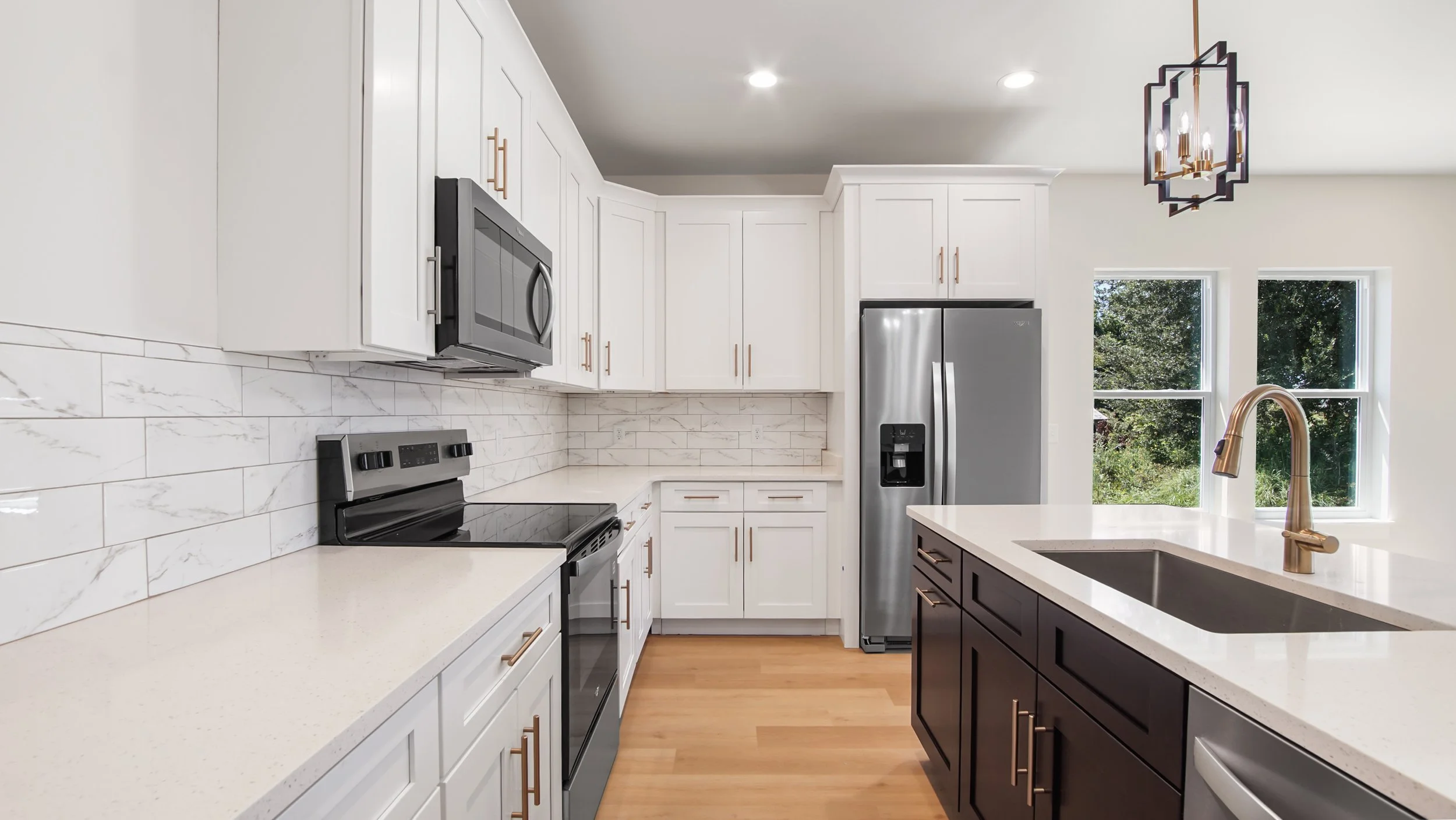 Modern kitchen with white cabinets, a black and stainless steel refrigerator, a built-in microwave, black stove, white countertops, and a kitchen island with dark wood cabinets and a marble sink. Two windows show greenery outside, and a contemporary geometric light fixture hangs from the ceiling.