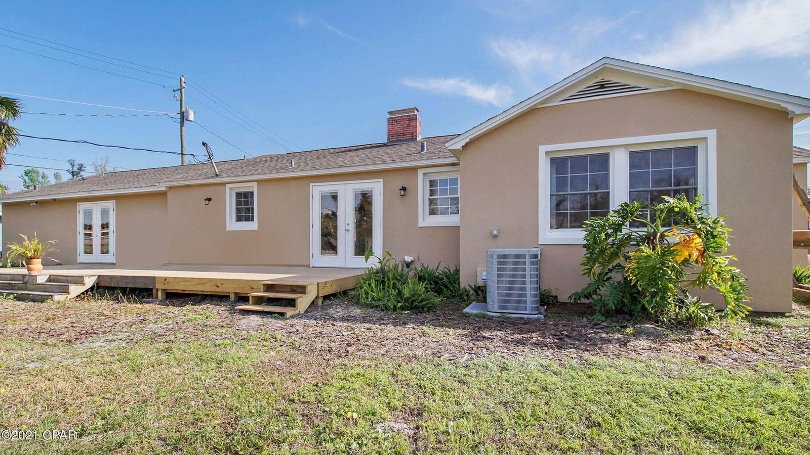 Backyard of a beige house with white trim, a wooden deck with stairs, a potted plant, and some bushes, under a blue sky.