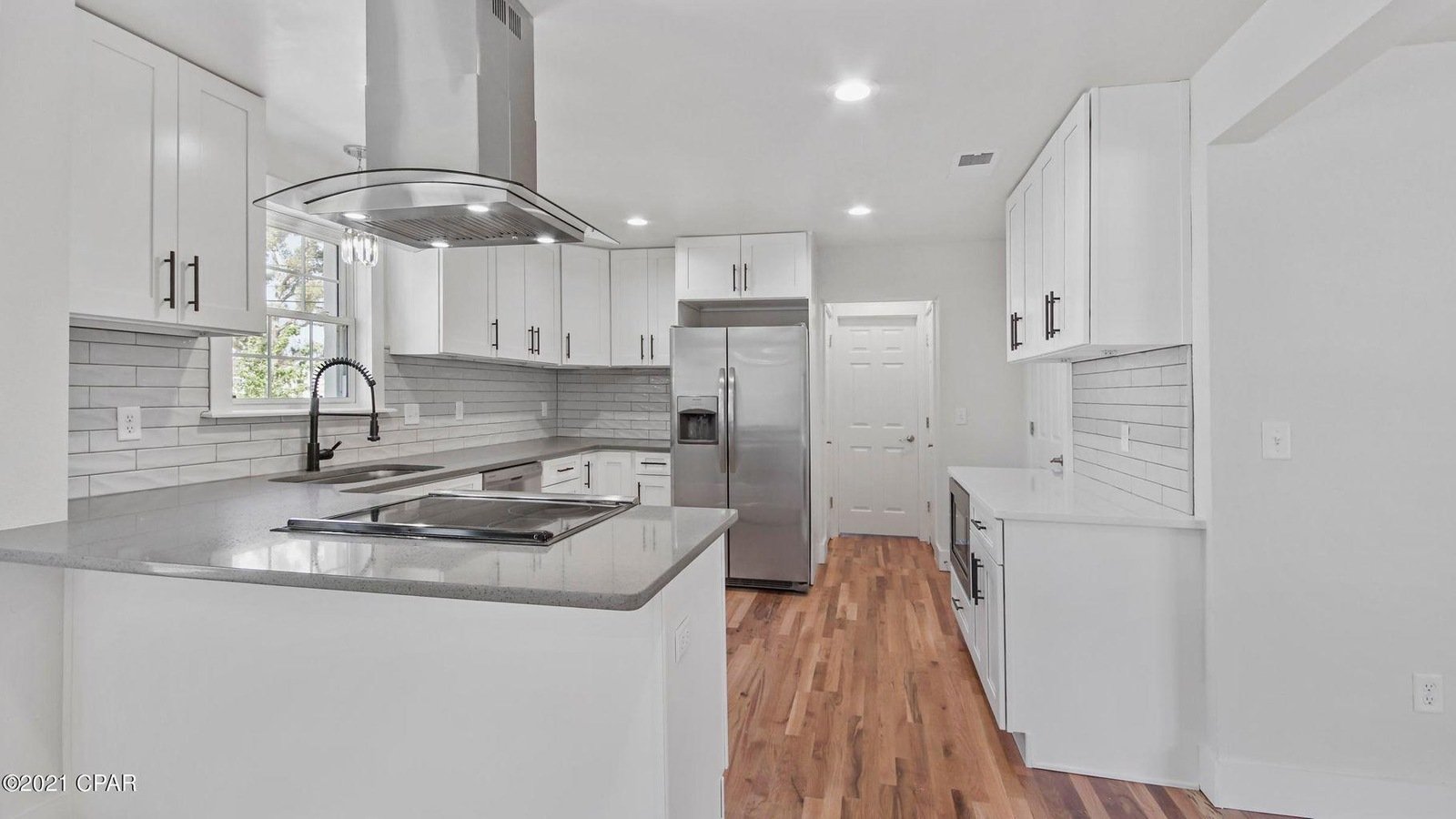 A modern kitchen with white cabinets, gray countertops, a stainless steel refrigerator, wood flooring, a black sink faucet, and a window over the sink.