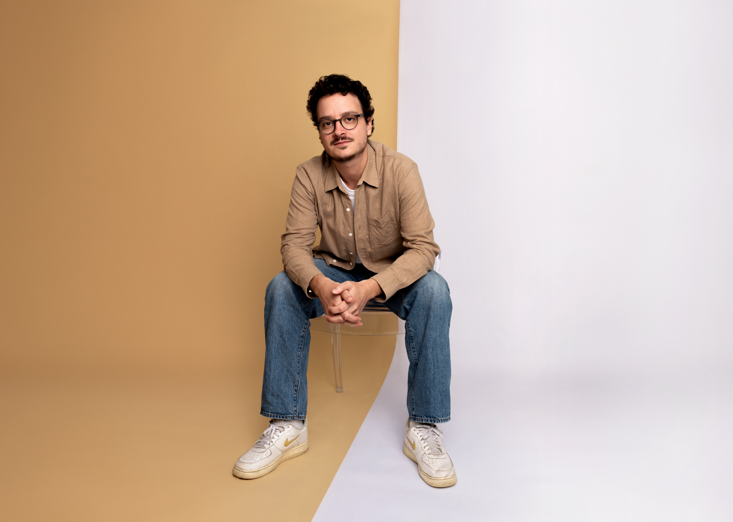 Jeune homme assis sur une chaise, vêtu d'une chemise beige, jeans, baskets blanches, avec fond plat moitié beige moitié blanc.