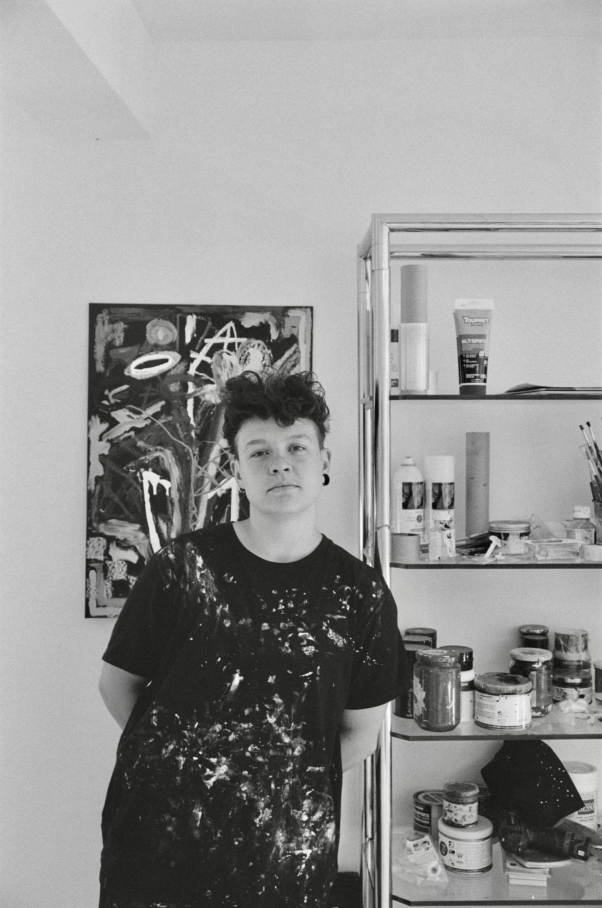 Jeune artiste debout dans un studio, portant un t-shirt taché de peinture, avec une peinture abstraite derrière lui et une étagère contenant des peintures et des fournitures d'art à côté.