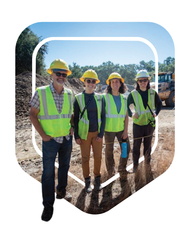 Four people at a construction site wearing yellow hard hats, high visibility safety vests, and casual work clothes, standing outdoors on a sunny day.