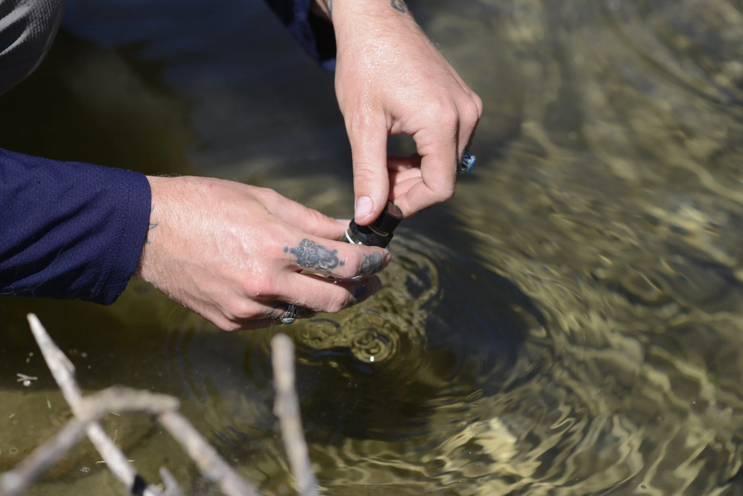 Person holding a small bottle or vial near water, possibly collecting a water sample.