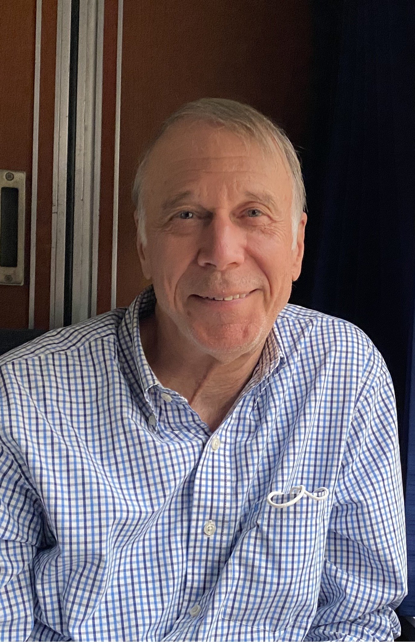 A young woman with curly hair, wearing glasses, a striped shirt, hoop earrings, and layered necklaces, smiling indoors.