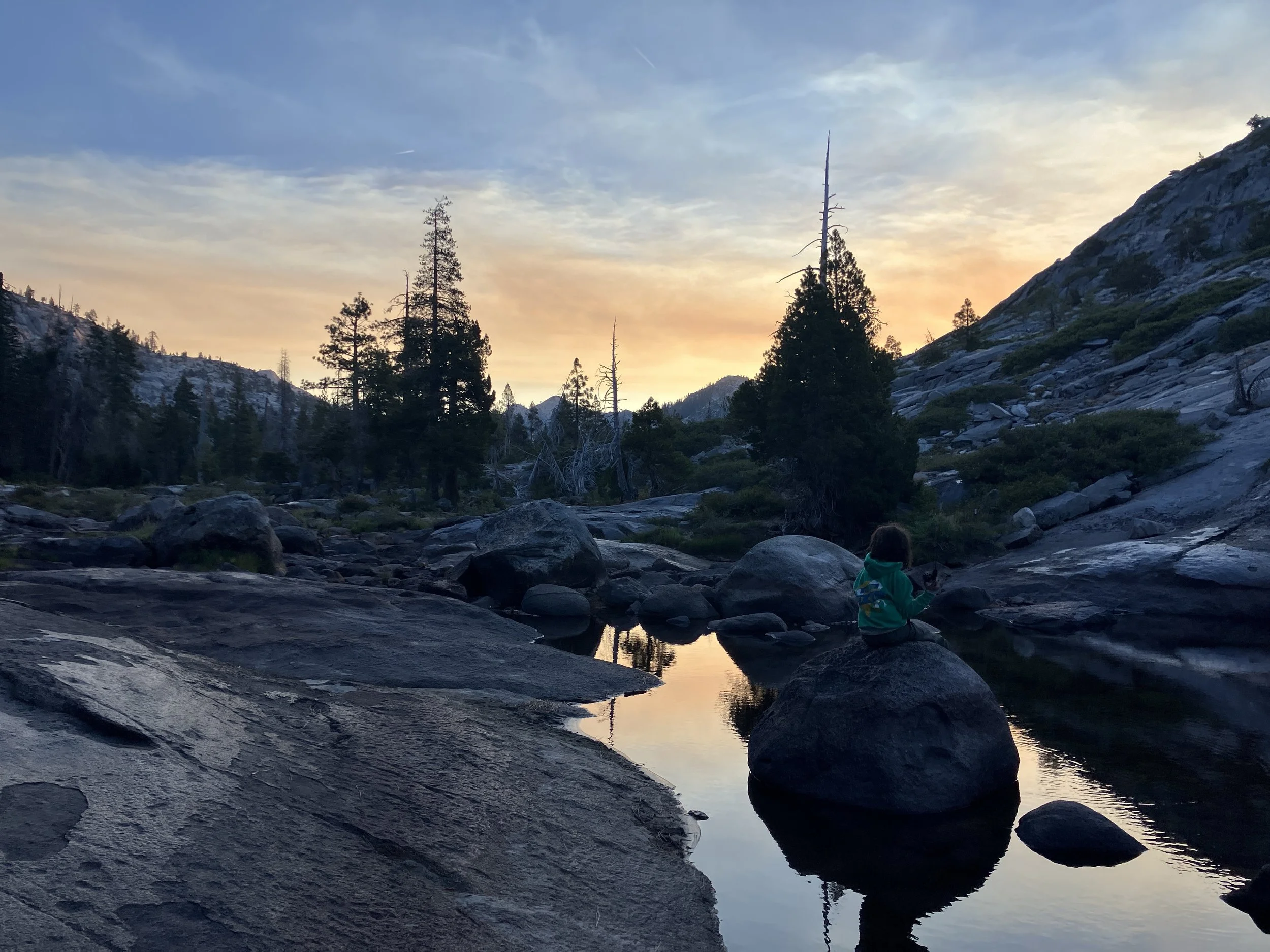 A person sitting on a rock in a mountain stream at sunset, with trees and rocky slopes in the background.