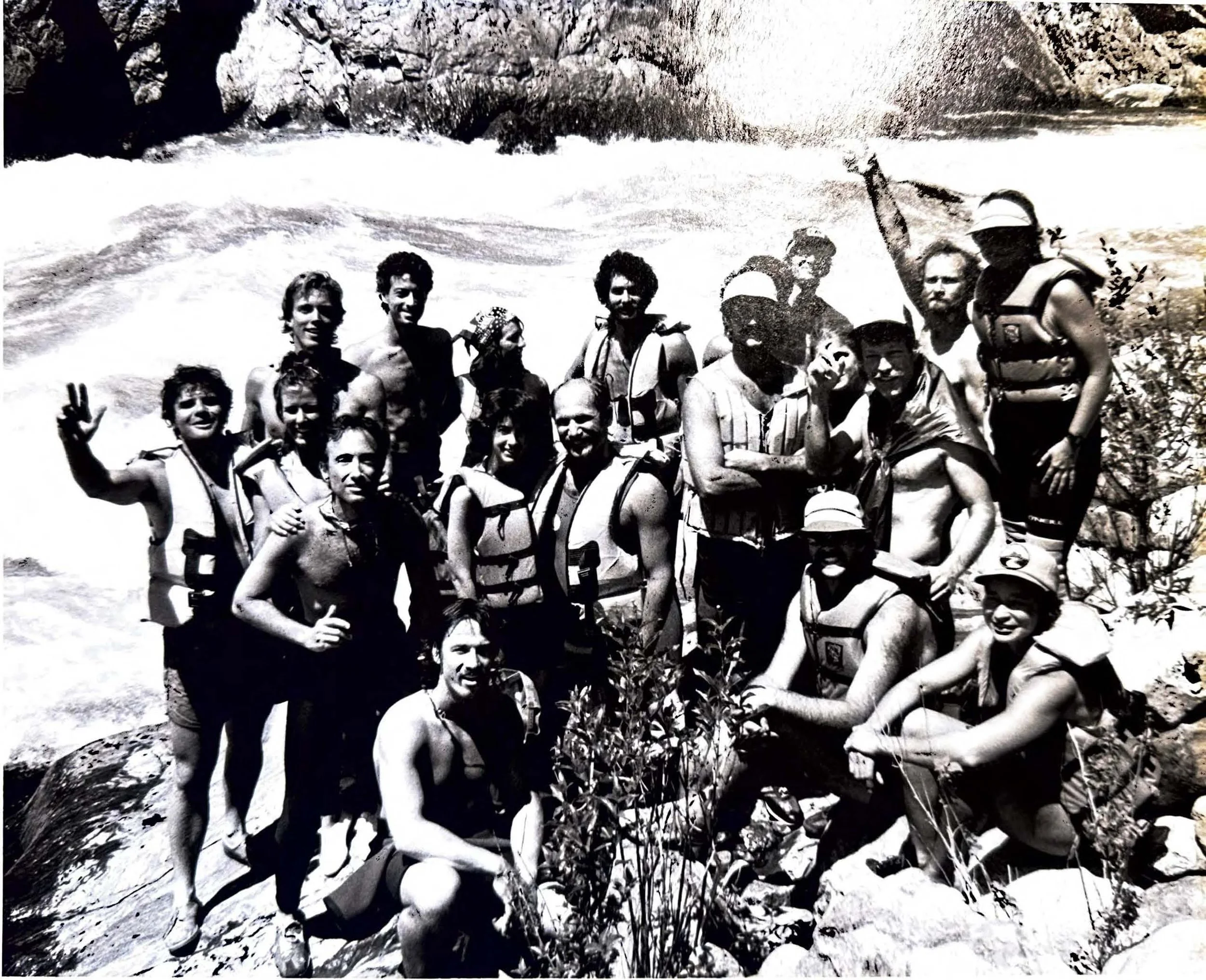 A group of people wearing life jackets and swimwear standing together on rocks near a river with rushing water.