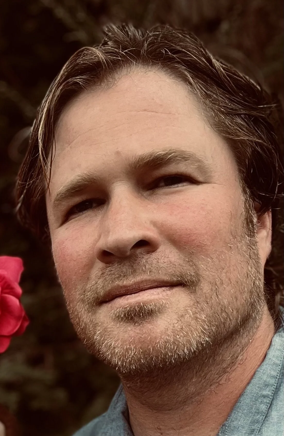 Close-up of a man with dark curly hair, clear glasses, and a beard, sitting indoors near a plant, looking to the side with a slight smile.