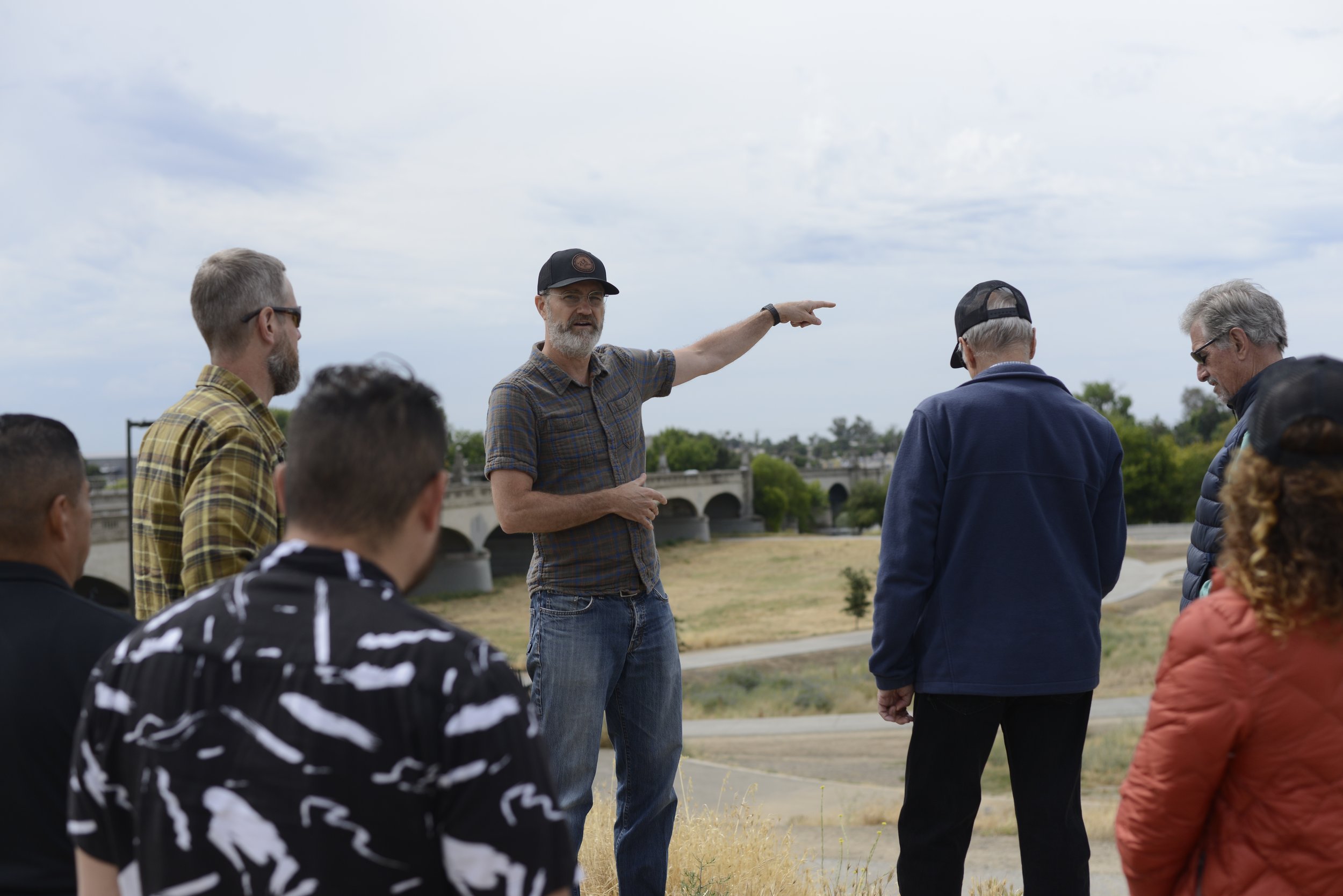 A man in a plaid shirt and cap is giving a tour or presentation to a group of people outdoors near a bridge. The group listens attentively, and the weather is cloudy.